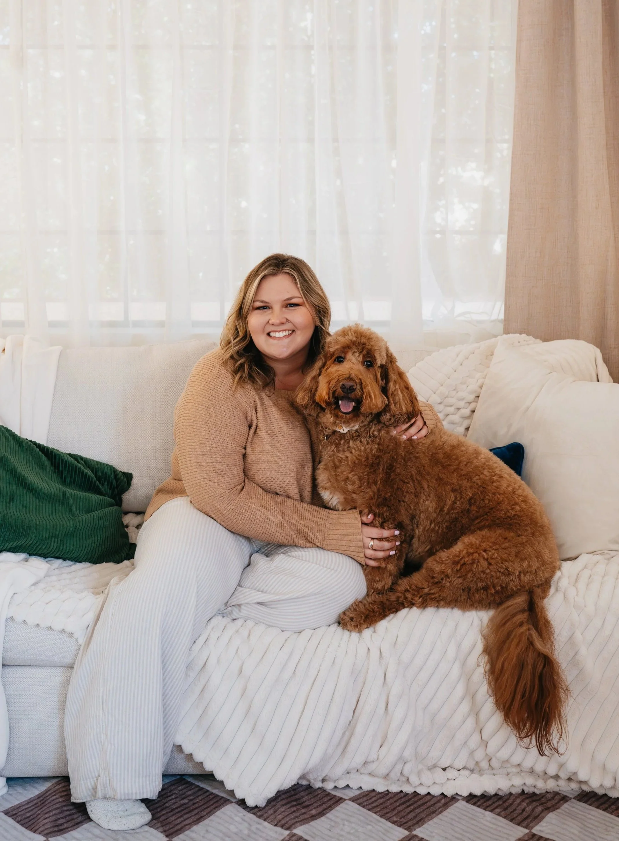 Smiling woman with shoulder-length blonde hair sitting on a cream-colored couch, hugging a large, fluffy goldendoodle dog with curly fur in a cozy living room with sheer curtains and throw pillows.