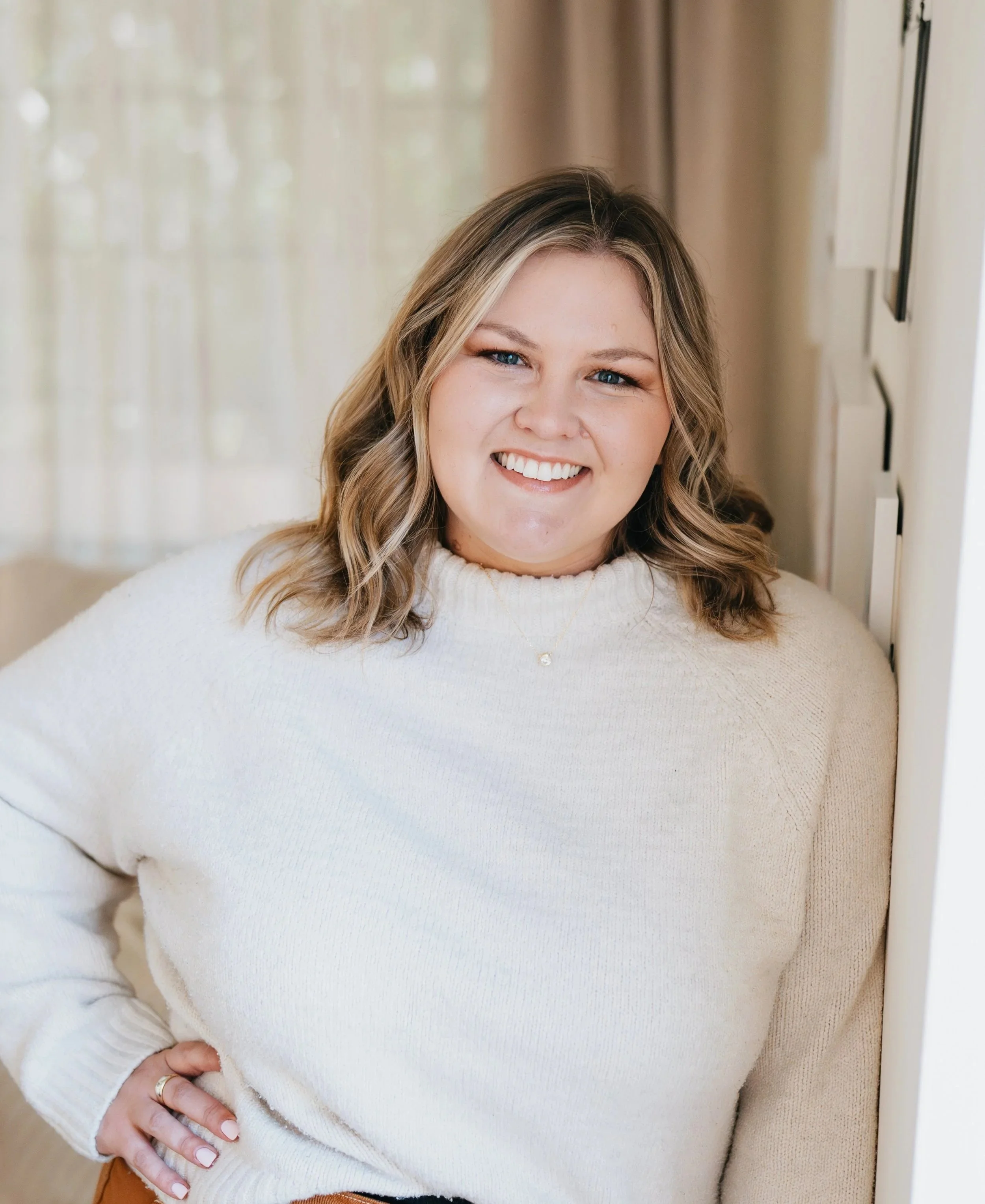 A woman with shoulder-length blond hair, smiling, wearing a white sweater, standing indoors near a wall with a window and curtains in the background.