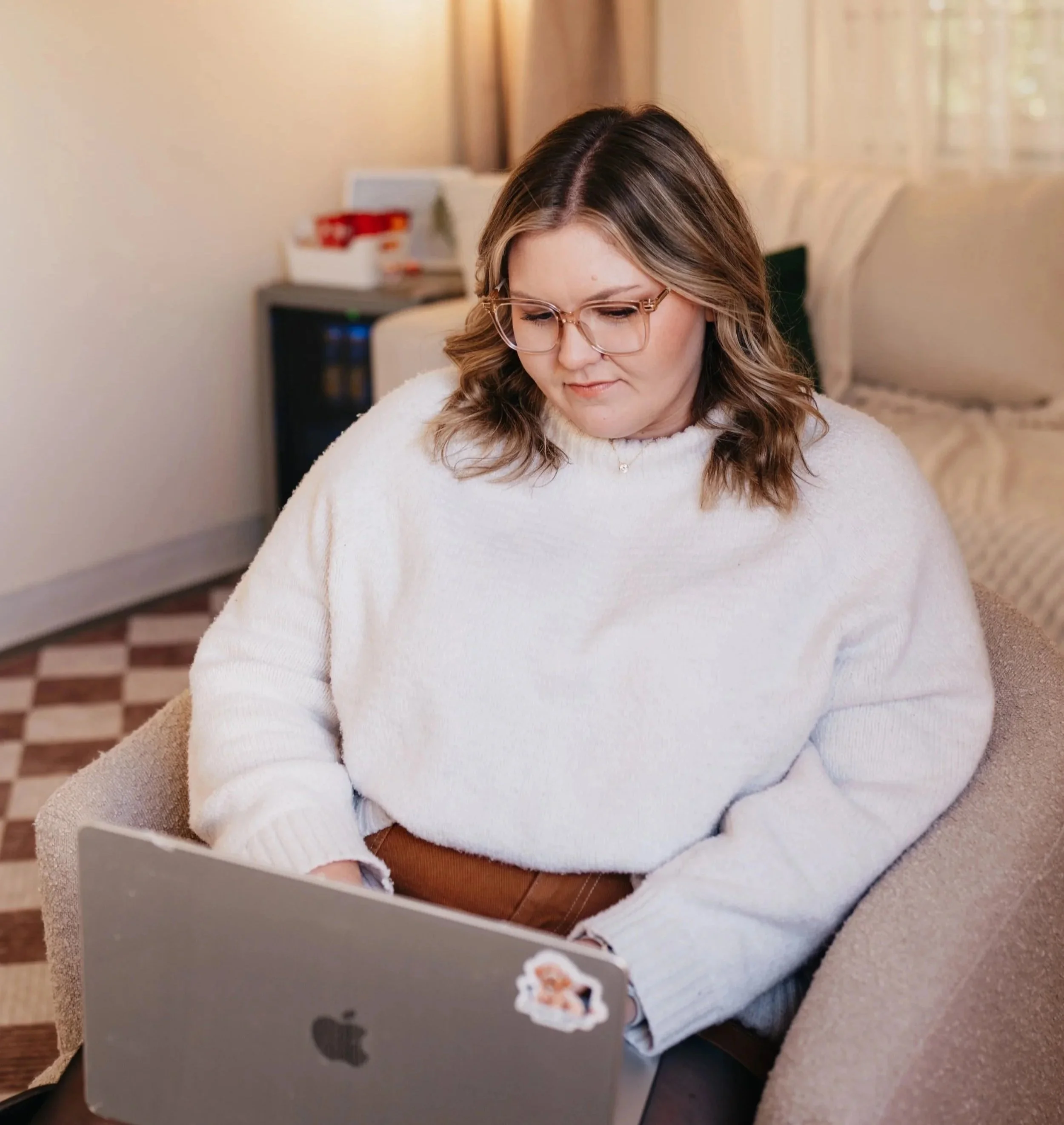 Woman with glasses sitting on a beige chair, working on a silver MacBook in a living room with a beige couch and patterned floor.