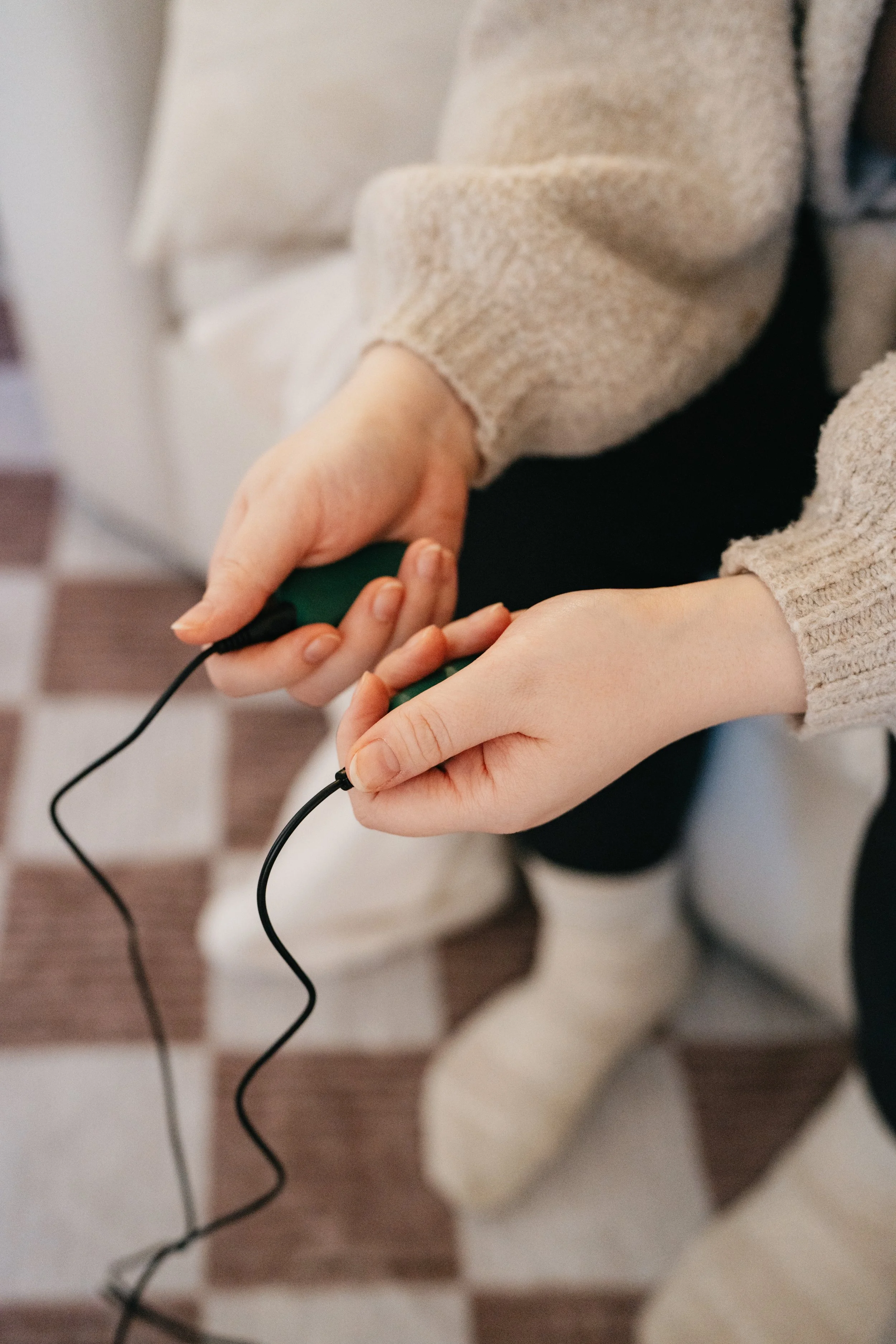 Person holding a green gaming controller with a black wire, sitting on a checkered wooden floor.