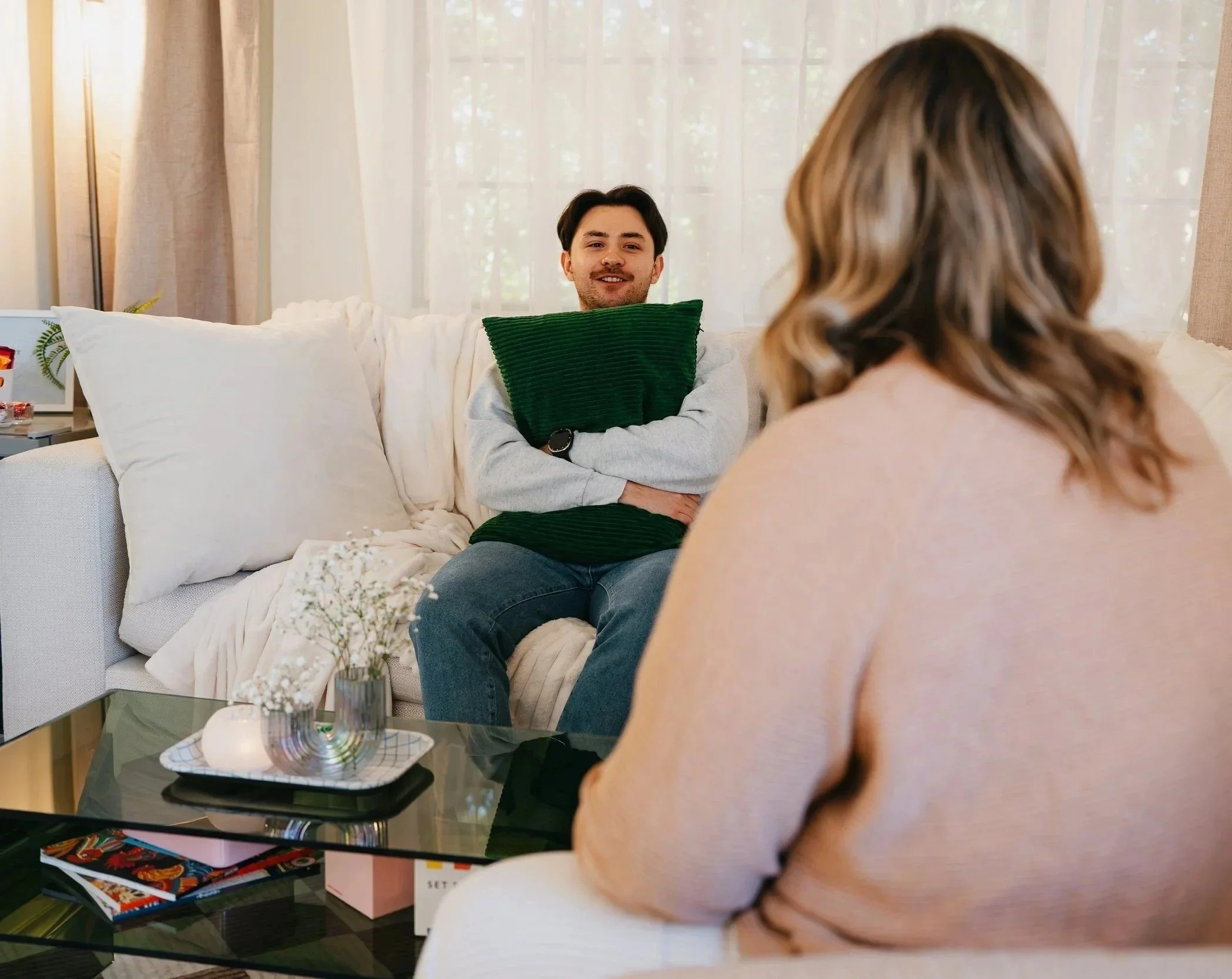 A woman and a man having a conversation in a living room, with the man sitting on a sofa holding a green pillow and smiling.