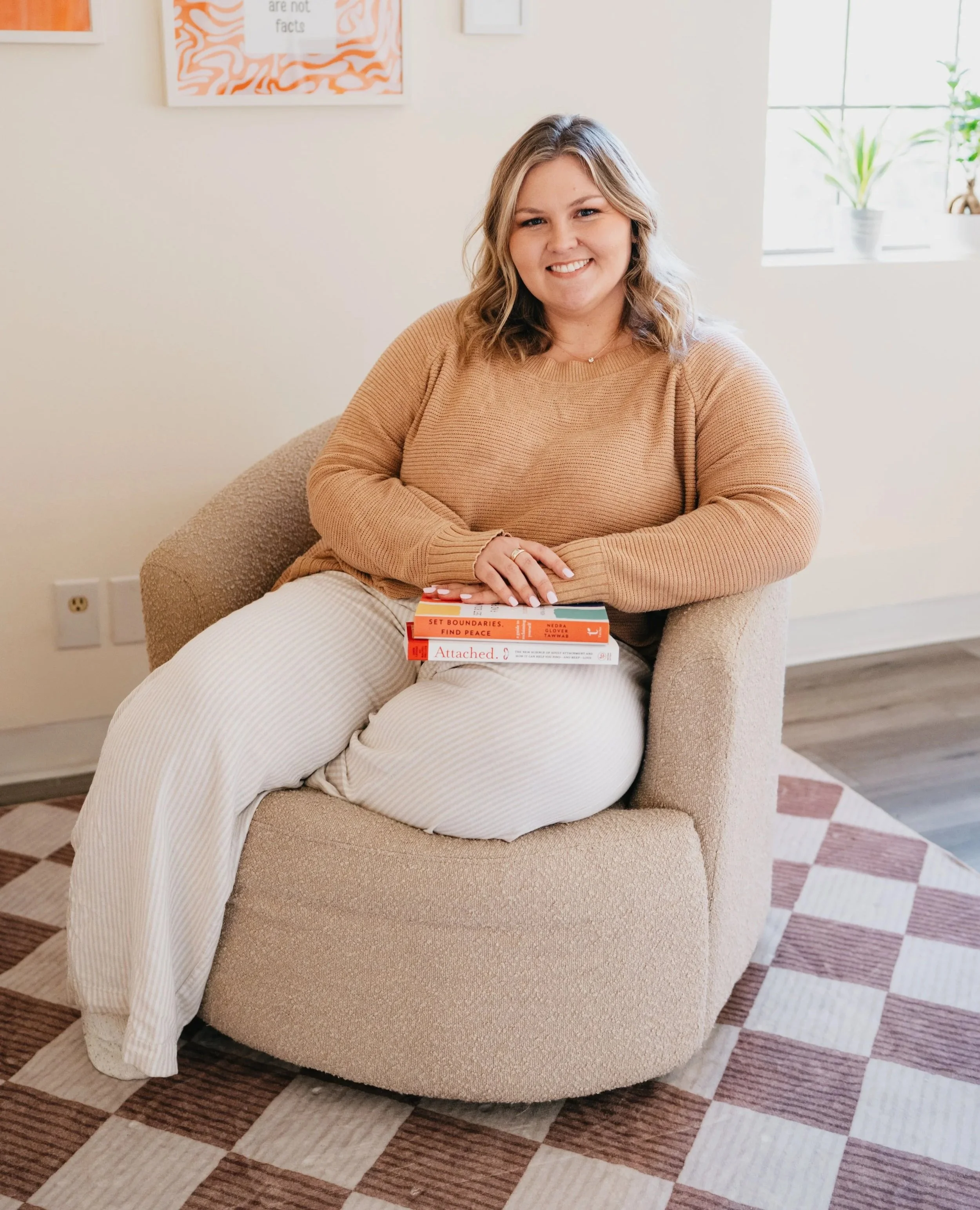 A woman with shoulder-length blonde hair sitting on a beige armchair indoors, holding two books on her lap called "Attached" and "Set Boundaries, Find Peace", and smiling at the camera. There is a window with a plant in the background.