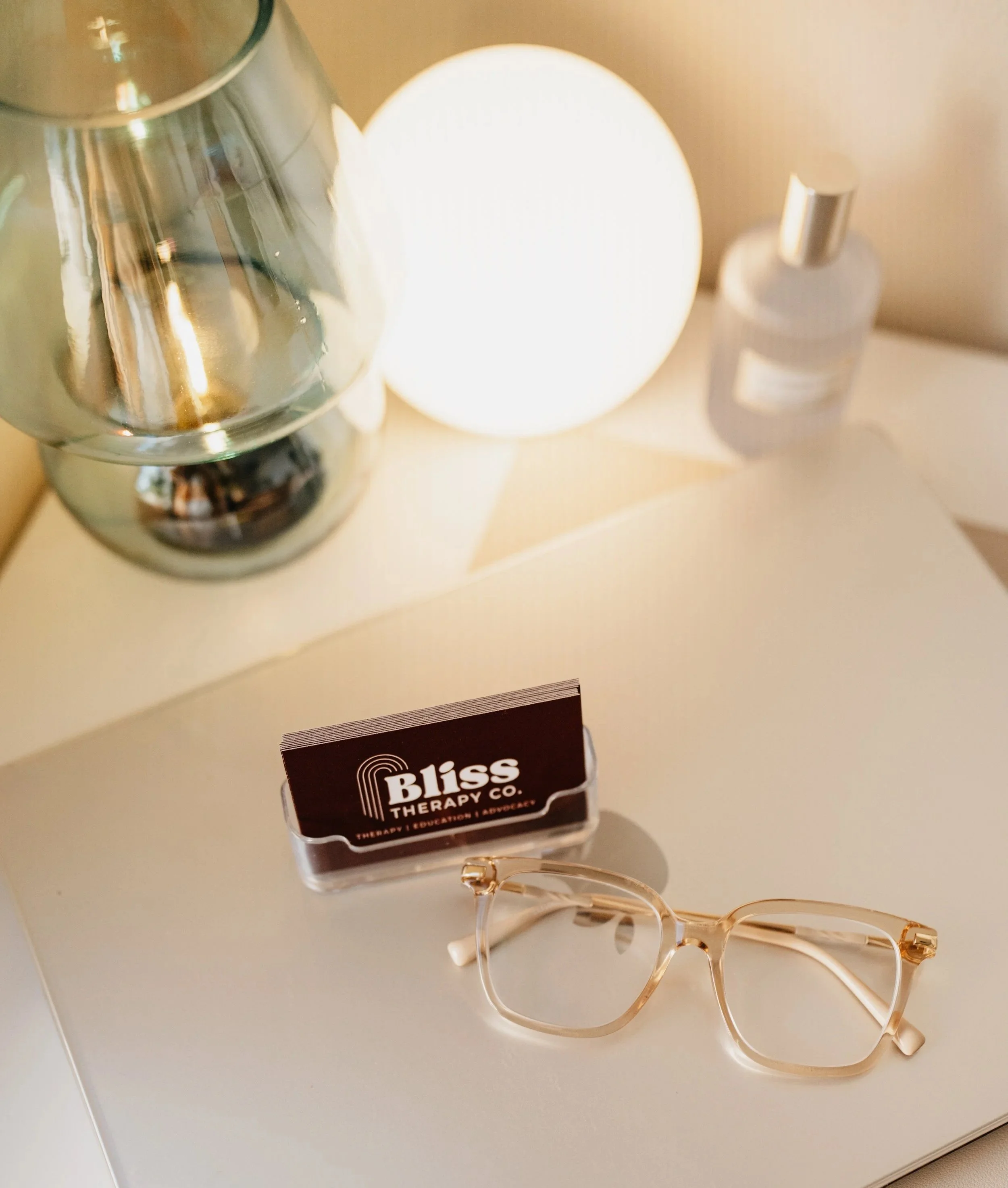 A pair of clear eyeglasses, a business card for Bliss Therapy Co., a lamp, a decorative silver vase, and a small white spray bottle on a white surface.