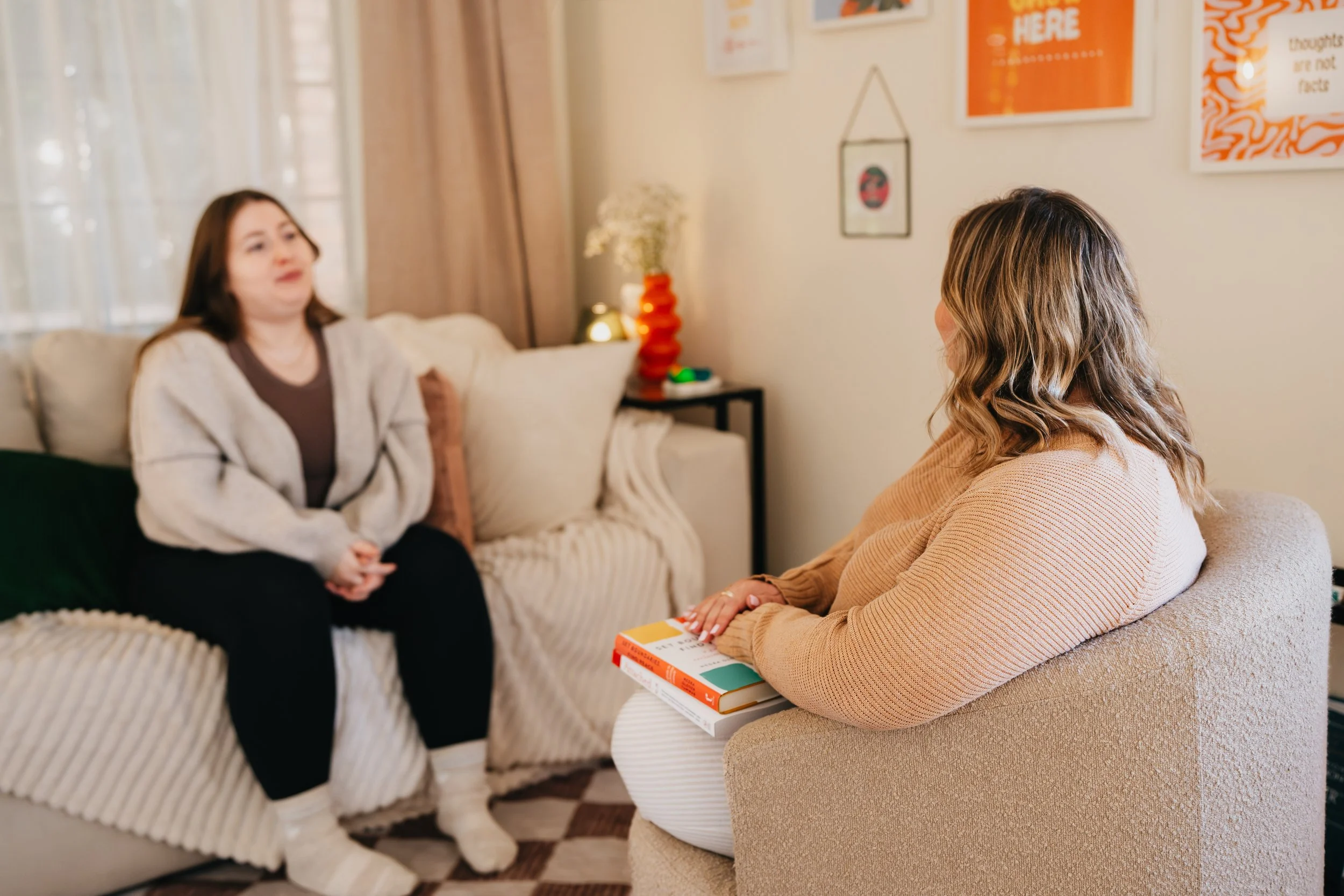 Two women having a conversation in a cozy living room, one sitting on a sofa with beige throw pillows and the other on an armchair holding a book, with wall art and a lamp in the background.