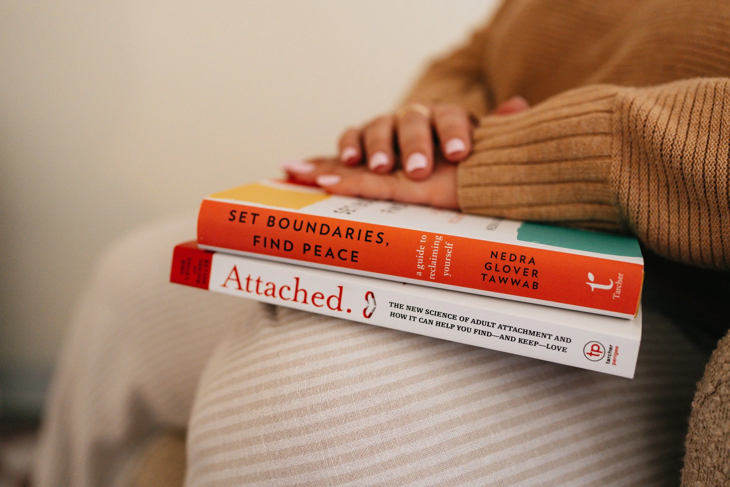 A person holding a book titled 'Set Boundaries, Find Peace' by Nedra Glover Tawwab, resting on their lap, with another book titled 'Attached' by therapist perspective visible underneath.