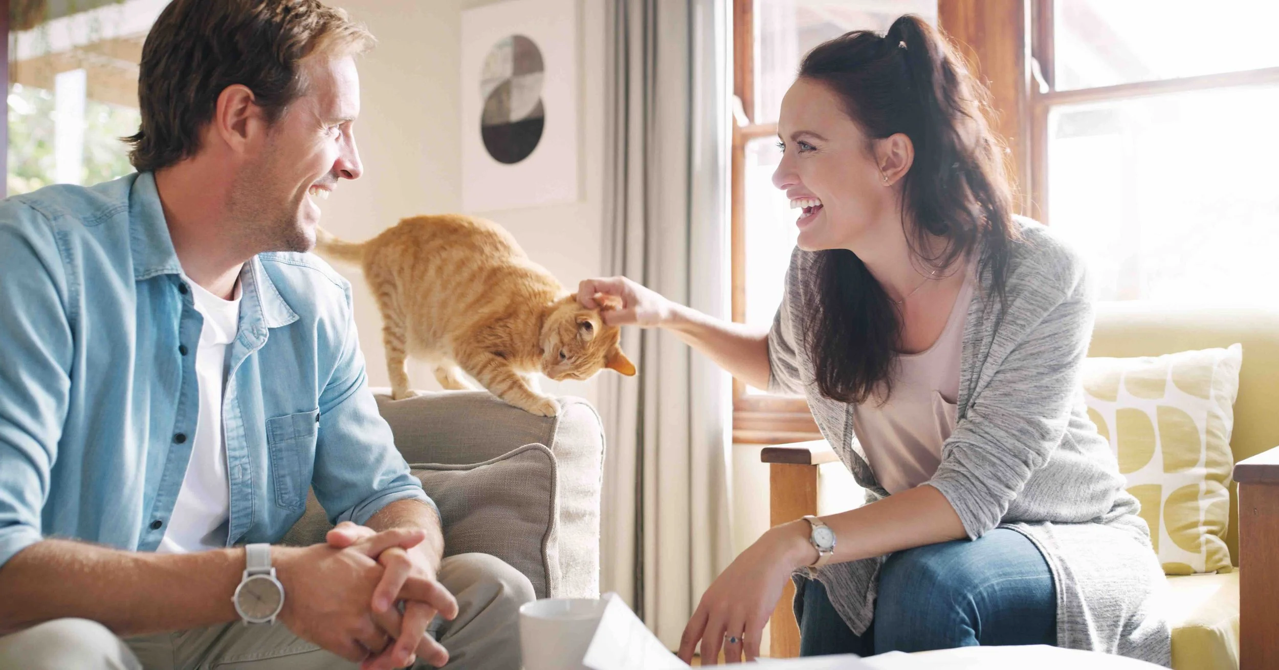 A man and woman laughing and playing with a ginger cat on a cozy living room couch. The woman is petting the cat while the man watches smiling.