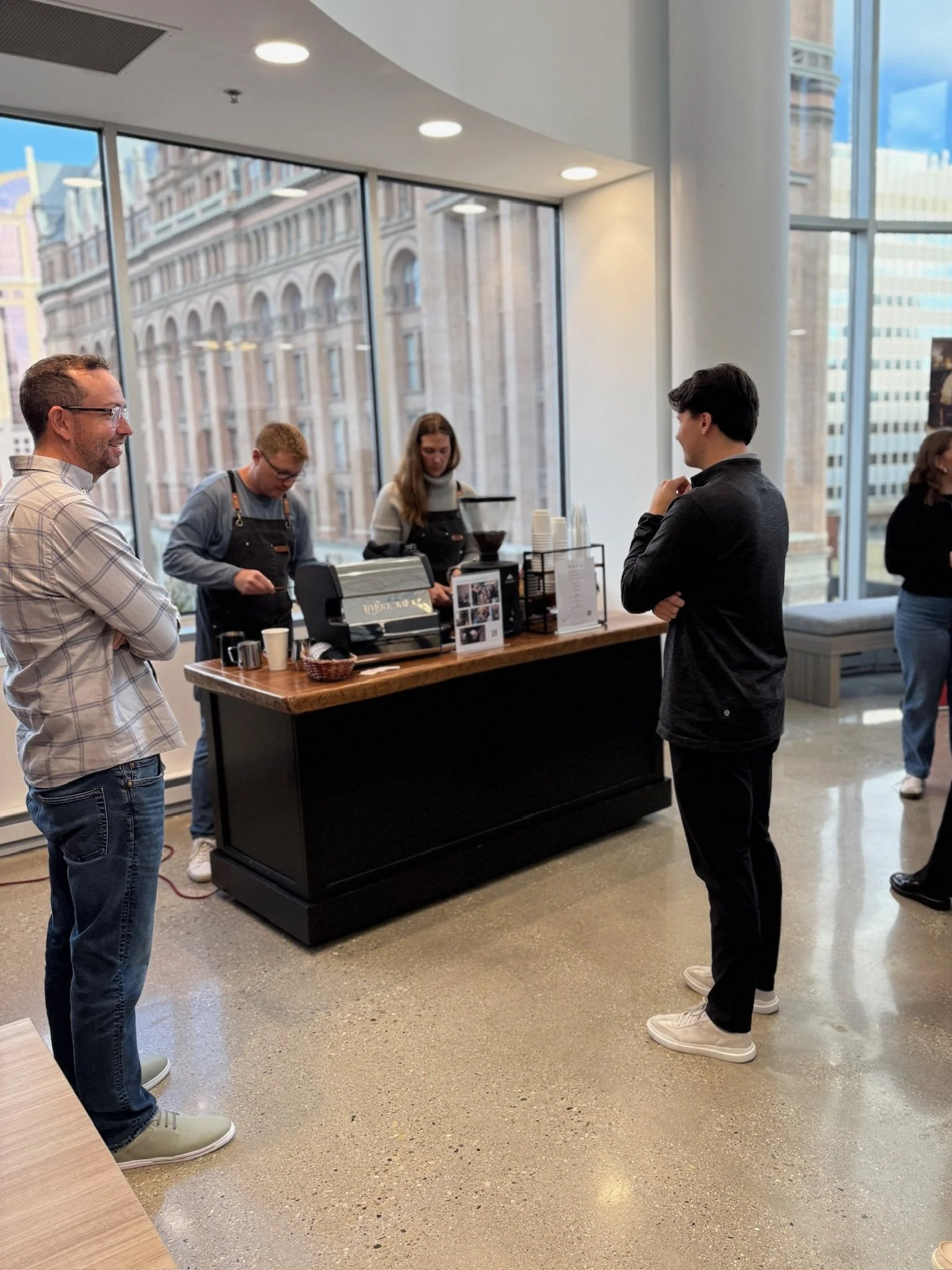 People standing at a coffee cart in a corporate office, with large windows showing Milwaukee buildings outside.