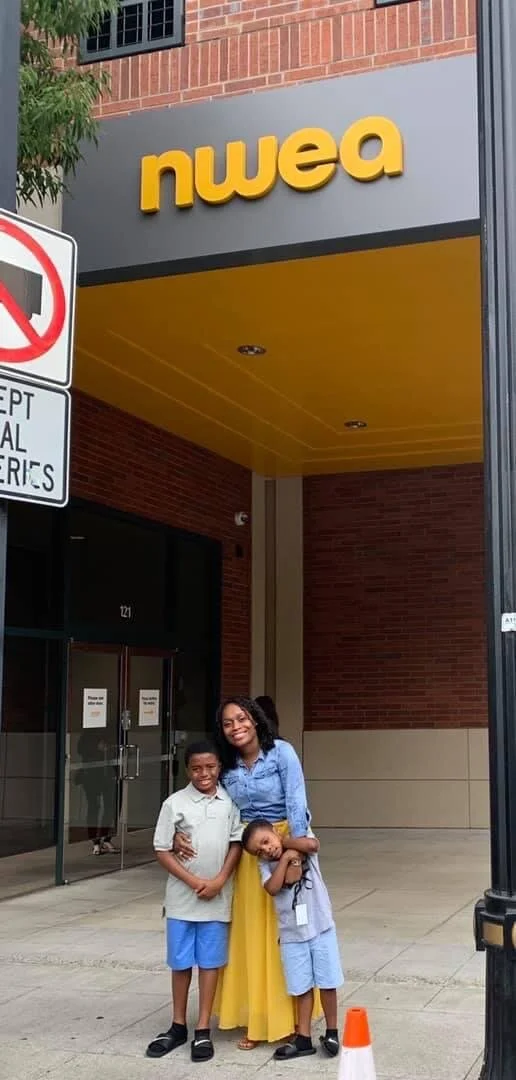 A woman and two young boys standing outside the entrance of a store called 'nwea,' smiling and hugging.