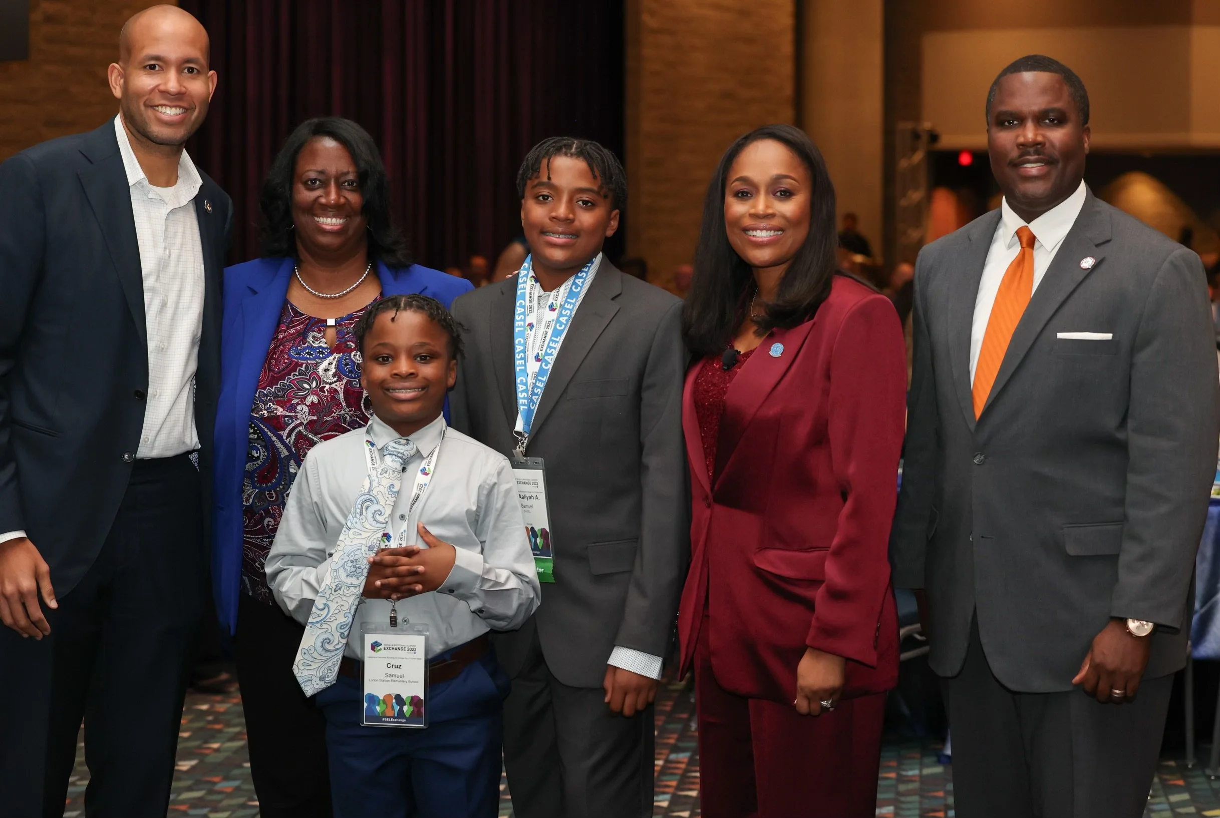 A group of seven diverse individuals, including children and adults, standing together at an indoor event, smiling at the camera.