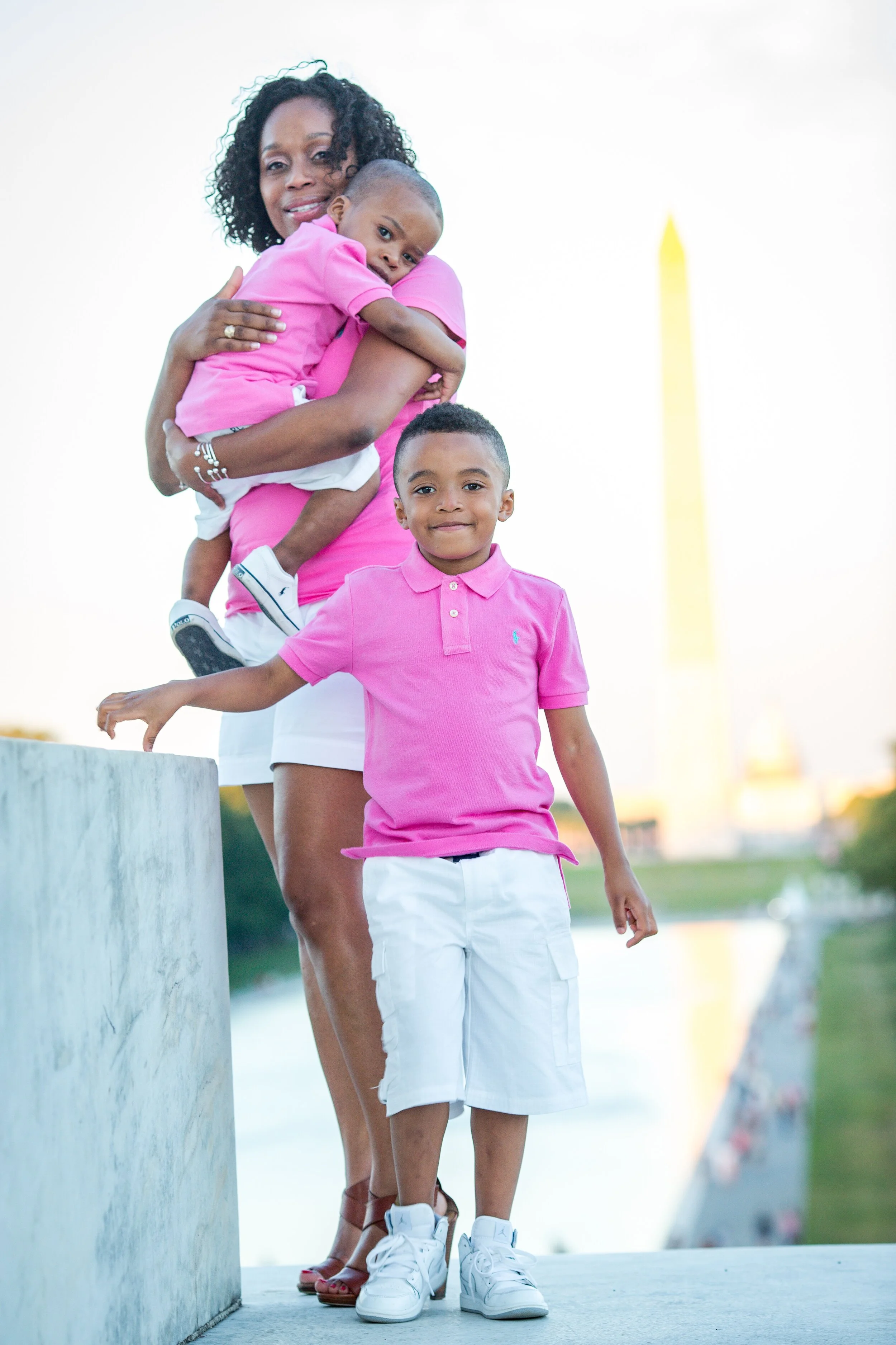 A woman holding a young child, standing with a young boy outdoors near the Washington Monument, with a river and a park in the background during sunset.