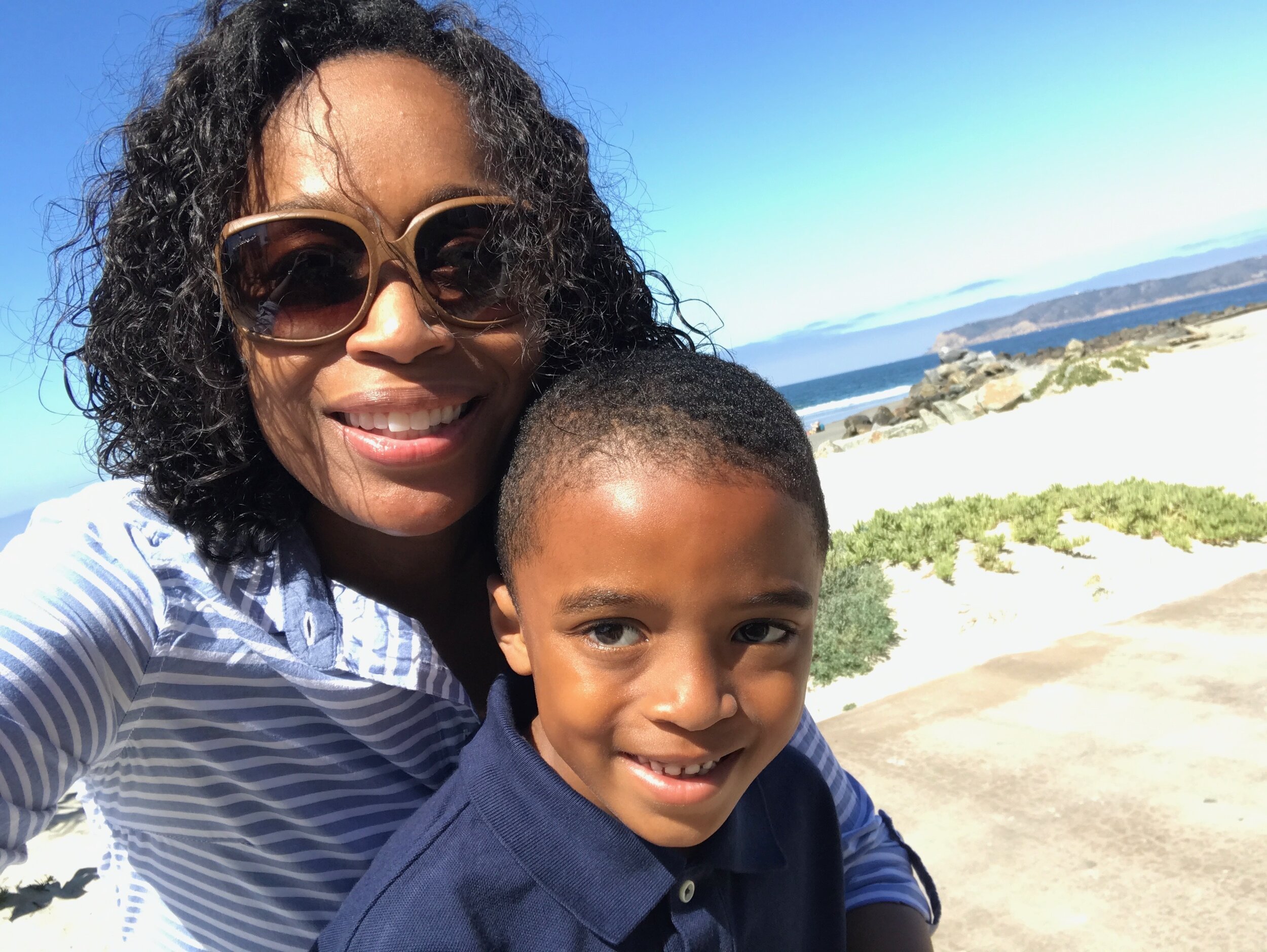 A woman and young boy taking a selfie at the beach with ocean and rocky coastline in the background. The woman has curly hair, is wearing large sunglasses, and a striped shirt. The boy has short hair and is wearing a navy blue polo shirt.