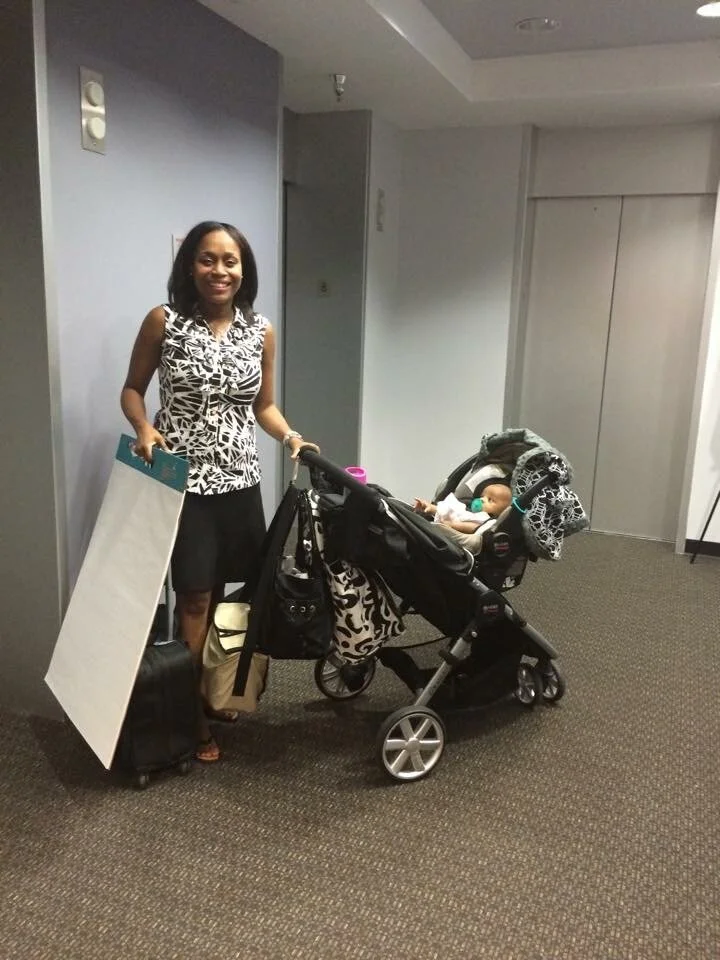 A woman standing next to a stroller with a baby, holding a large white and blue board, in an indoor hallway.