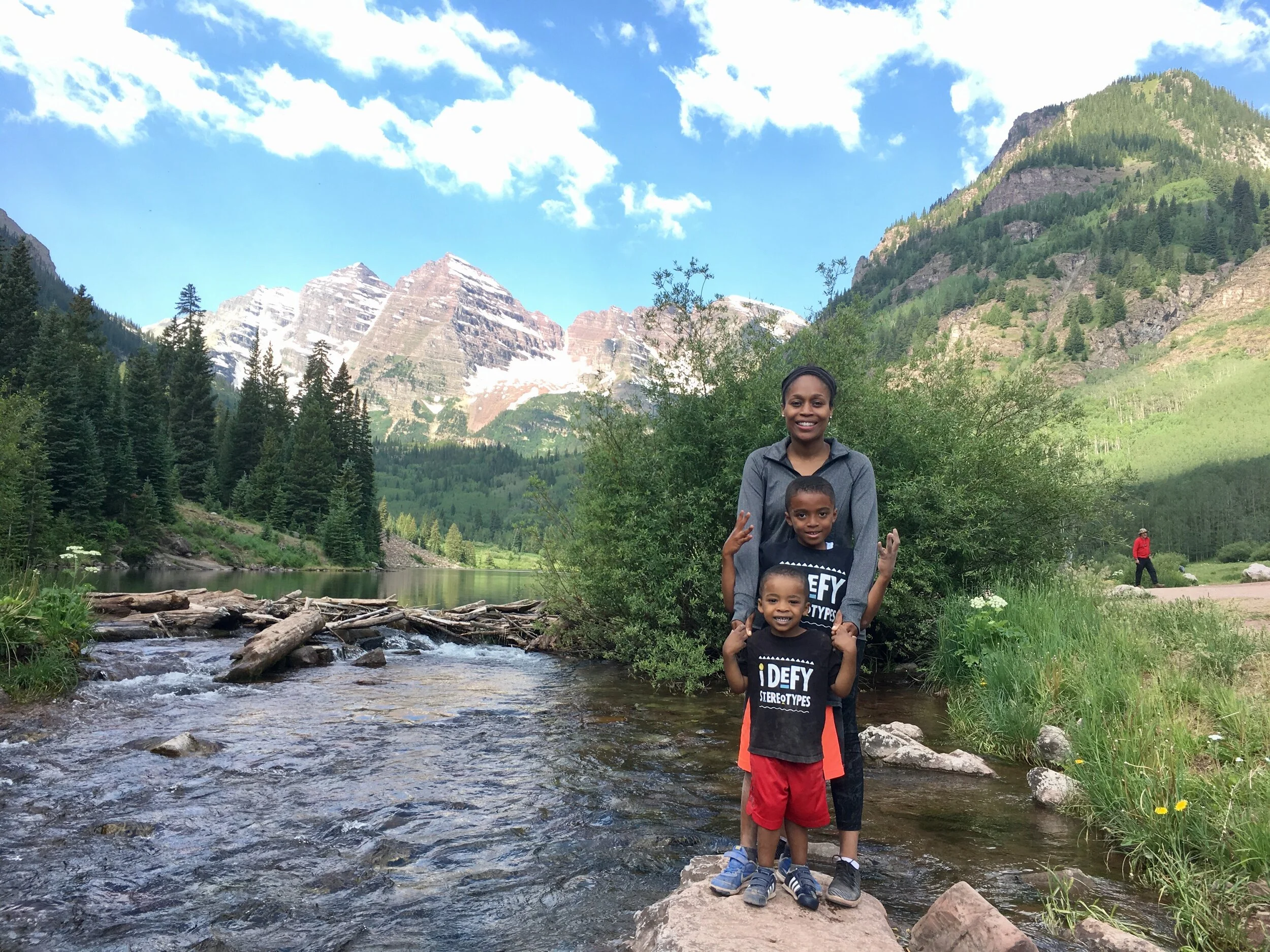 A woman and two young boys standing in a creek with mountains and a forest in the background.