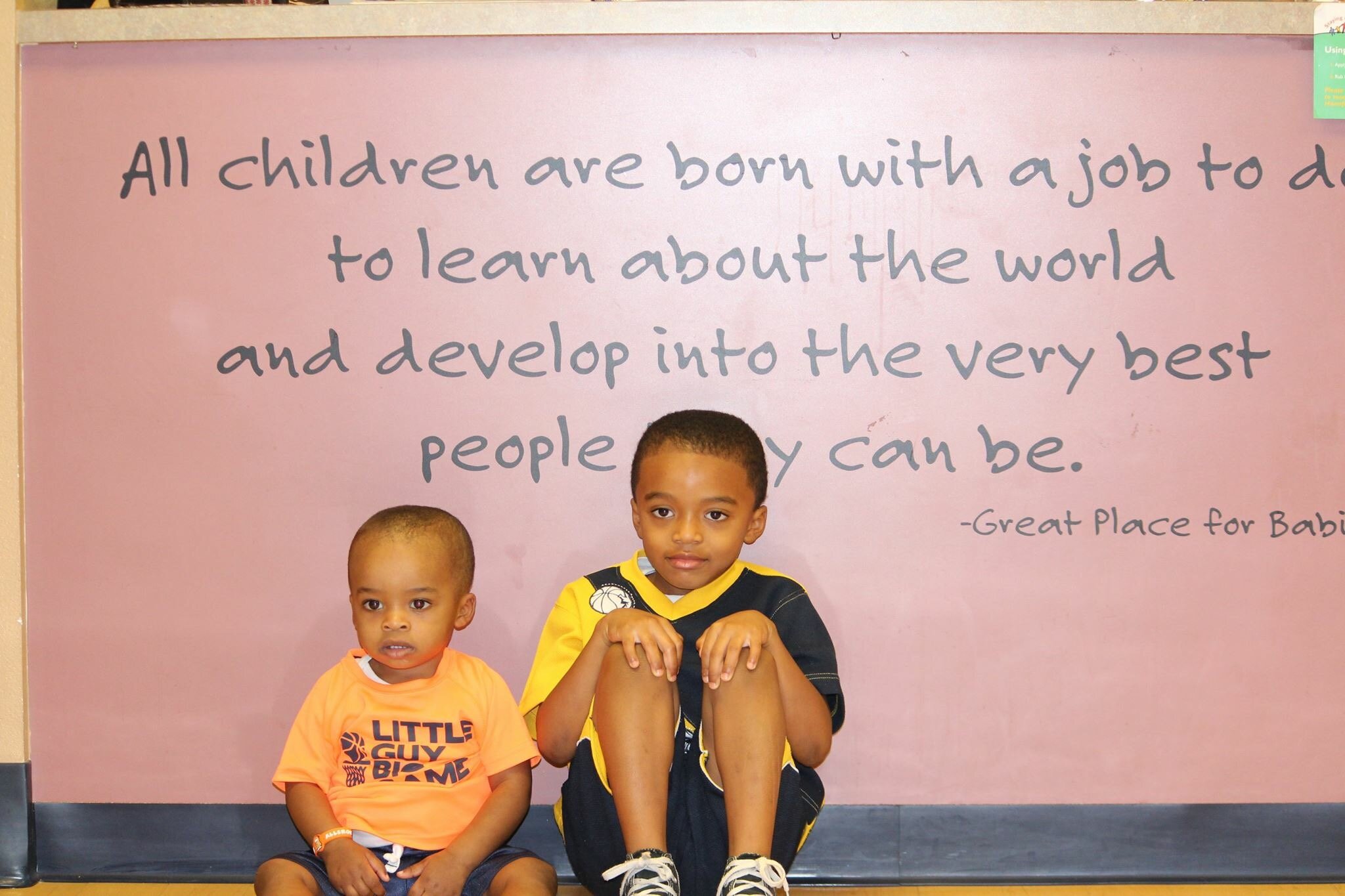 Two young boys sitting in front of a pink wall with a quote about children learning to understand the world.
