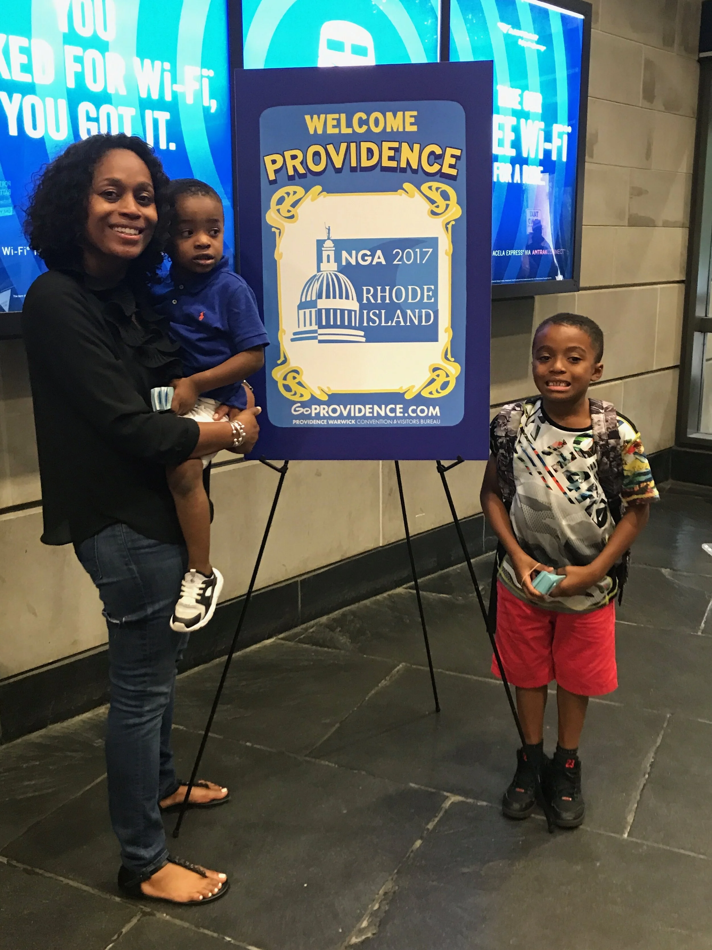 A woman holding a young boy standing next to a sign that says 'Welcome Providence, NGA 2017 Rhode Island', with a boy standing nearby holding a phone. They are in an indoor location with a digital screen in the background.