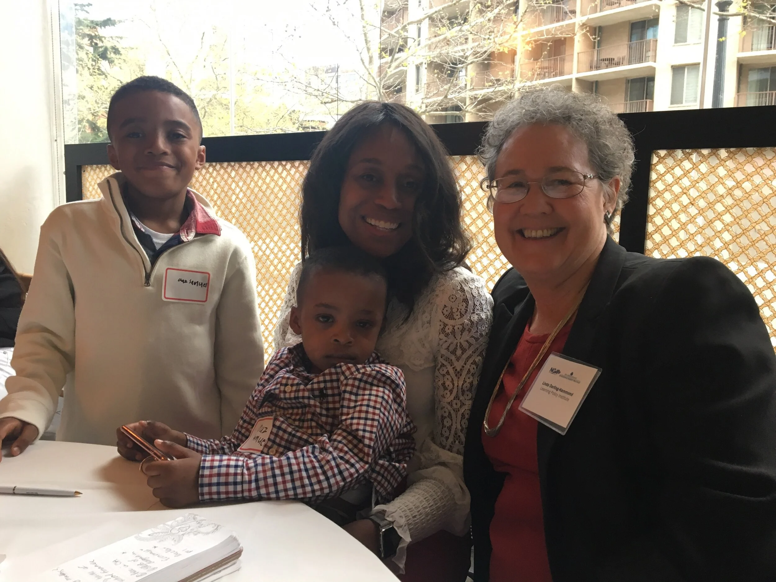 Four people, including two children and two women, posing together in a well-lit room near a window with trees and buildings outside.
