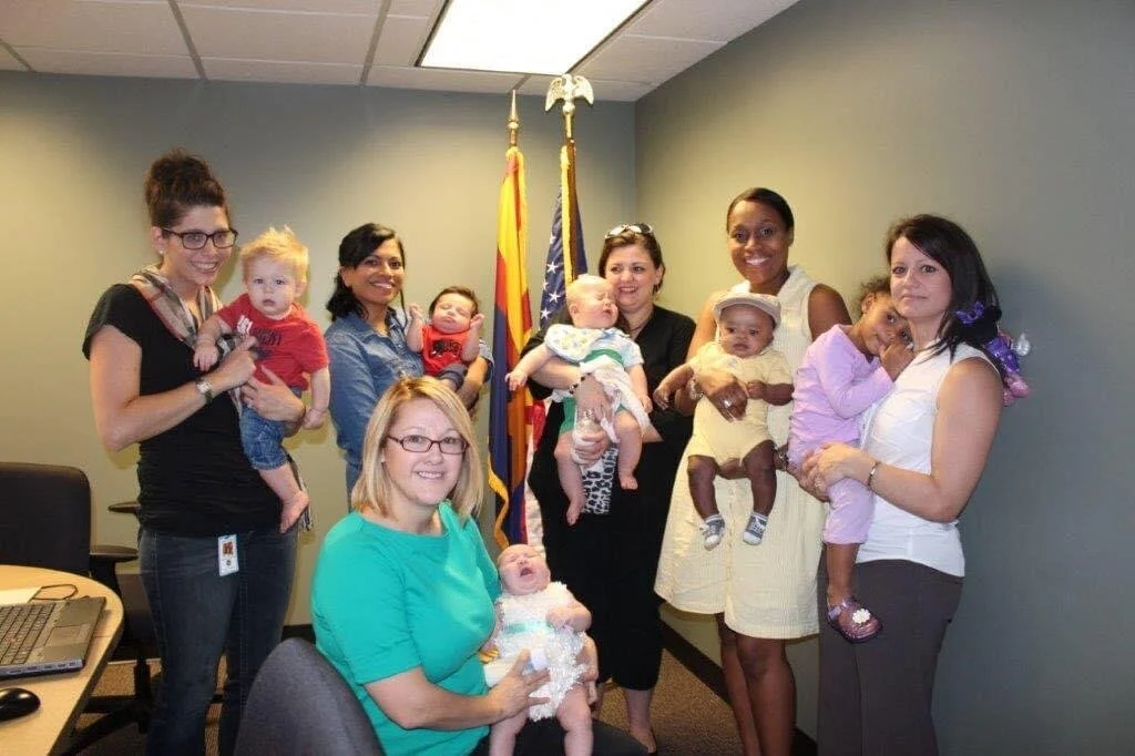 Group of eight women holding six children, with two flags in the background, in an office room.