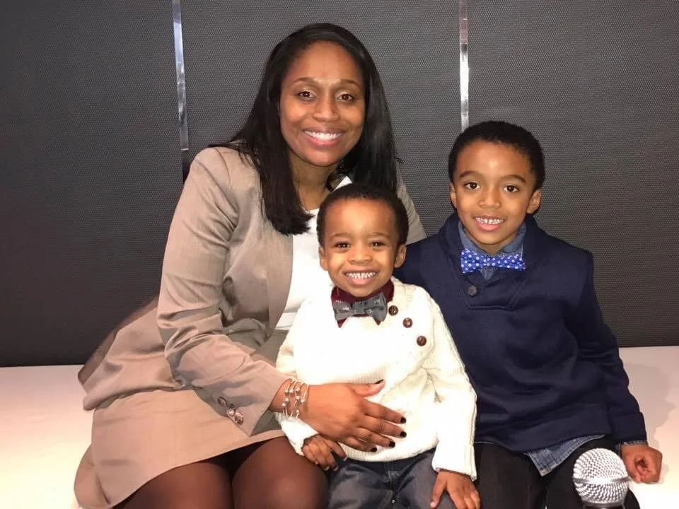 Smiling woman with two young boys, one wearing a white sweater and the other in a navy jacket with a bow tie, sitting together on a bench in an indoor setting.