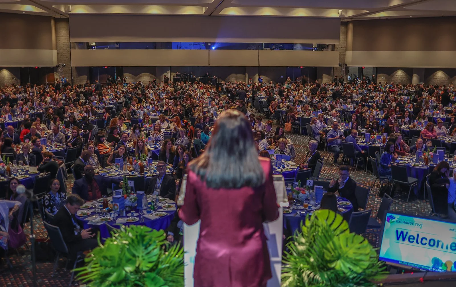 A woman with long gray hair wearing a maroon blazer speaks at a large conference or banquet in front of a seated audience in a ballroom. The audience consists of many people sitting at round tables decorated with floral centerpieces and table settings. There is a large screen to the right displaying a welcome message.