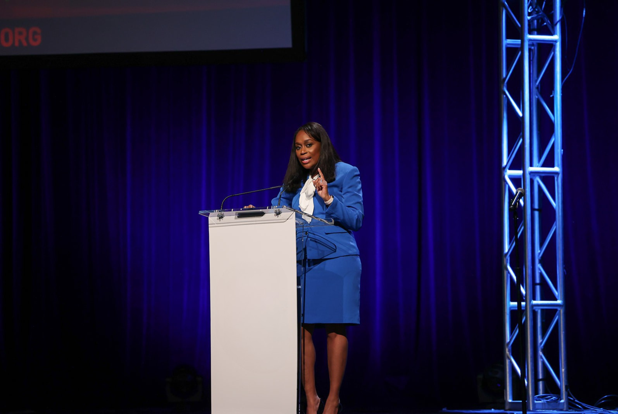 A woman in a blue suit is speaking at a podium on a stage with blue curtains and lighting.