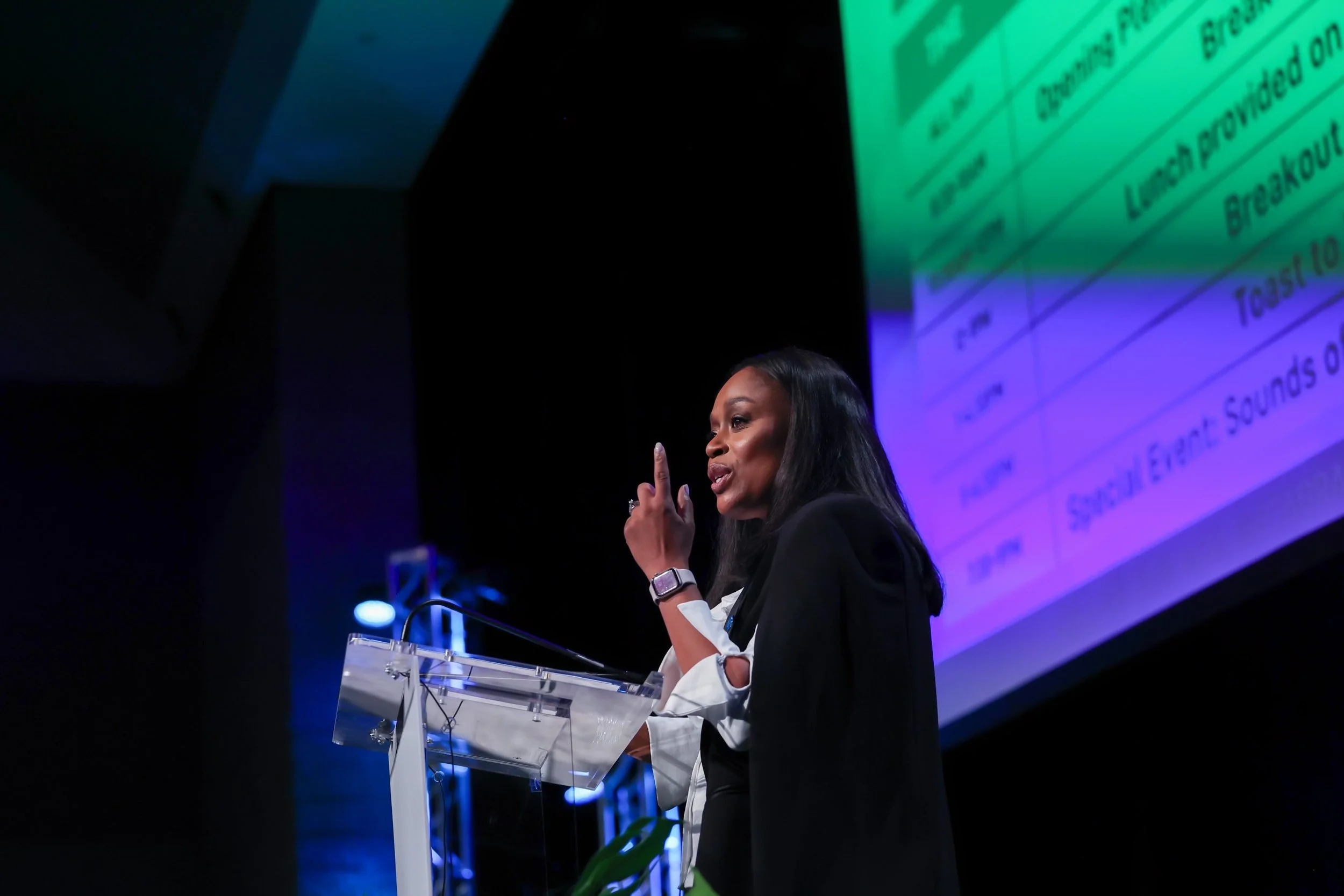 A woman in a black blazer and white blouse speaking at a podium with a microphone, gesturing with her right hand, in front of a large screen displaying a spreadsheet or table.