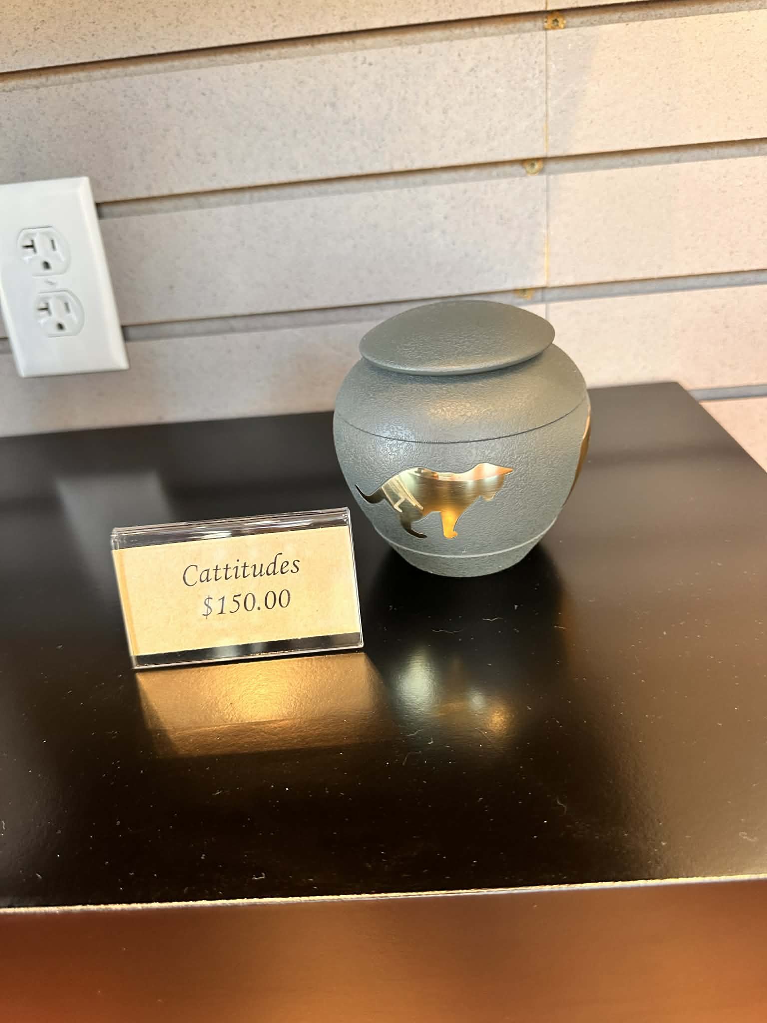 A small, decorative, spherical vase with cut-out shapes sits on a black surface; in front of it is a yellow price tag reading '$150.00', and a white electrical outlet is seen on the beige wall in the upper left corner.