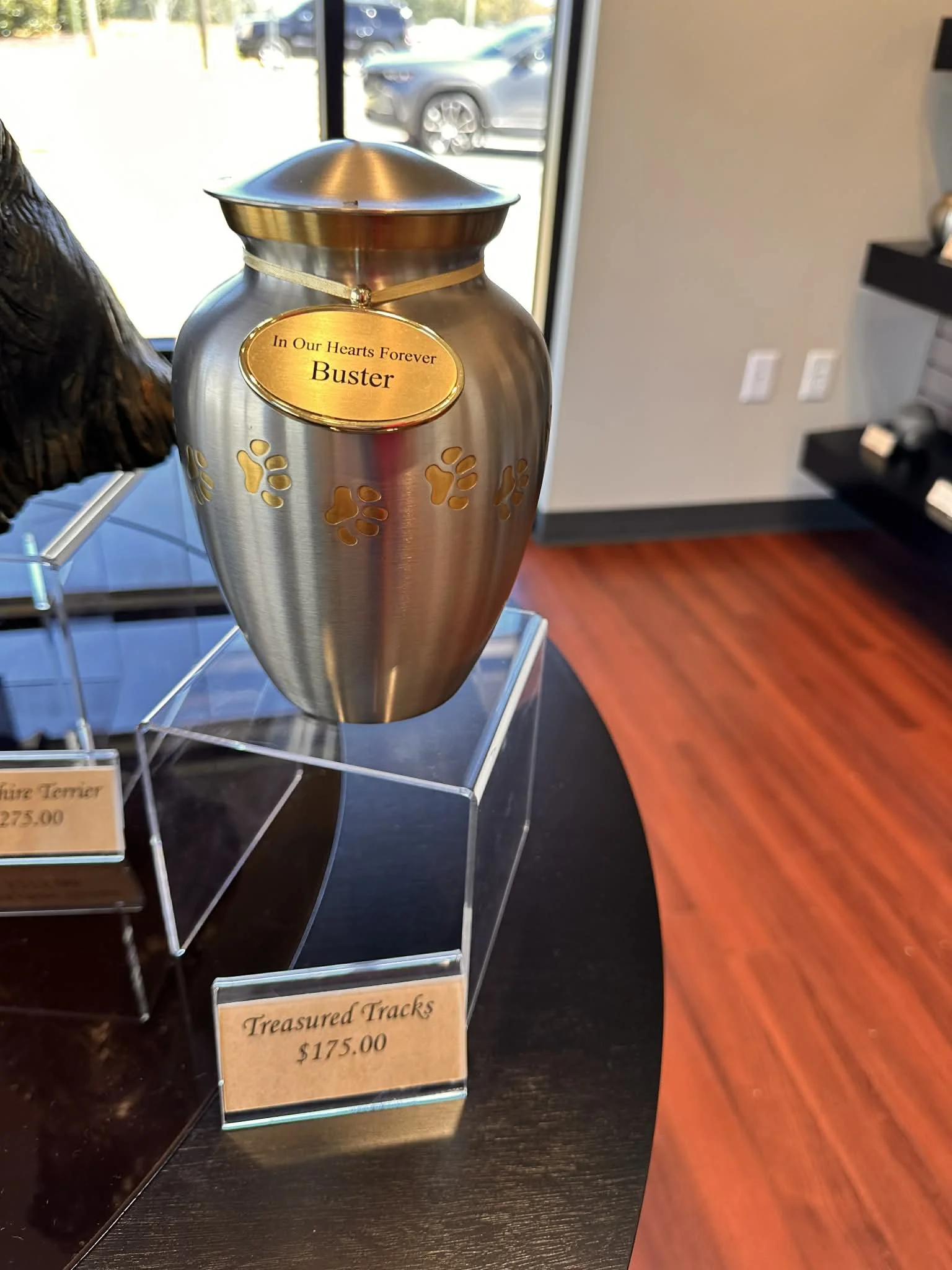 A metallic urn with paw print cutouts and a gold nameplate that reads 'In Our Hearts Forever Buster'.