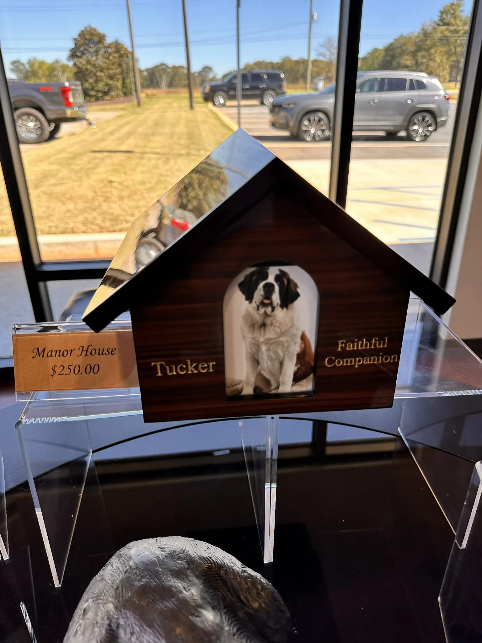 A small model doghouse labeled 'Manor House' priced at $250.00, with a photo of a puppy on the front and the words 'Trust & Faithful Companion.' The doghouse is displayed inside near a large window with parked cars and trees outside.