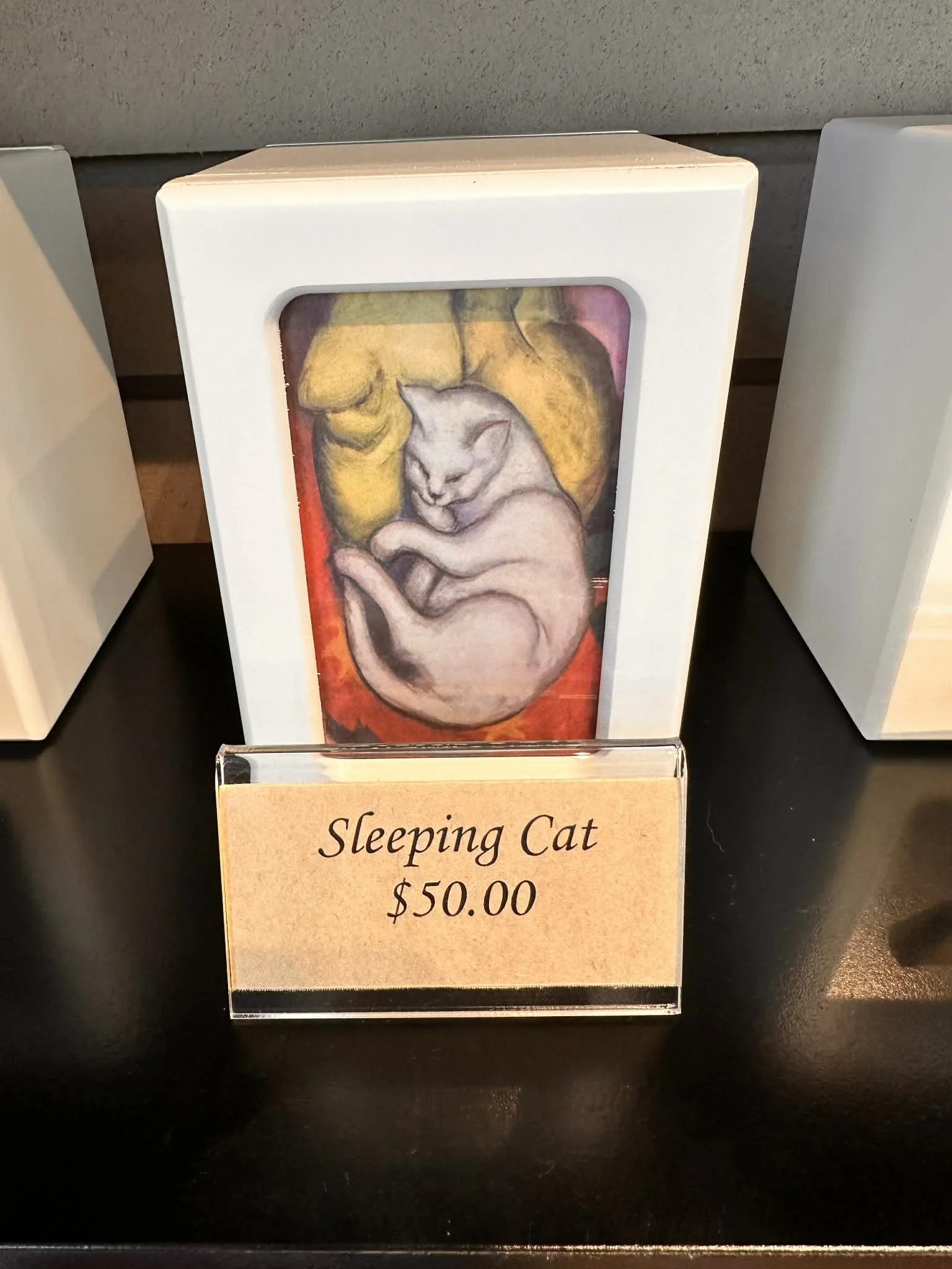A sign with a painting of a sleeping cat inside a white frame, and a label below that reads "Sleeping Cat $50.00".