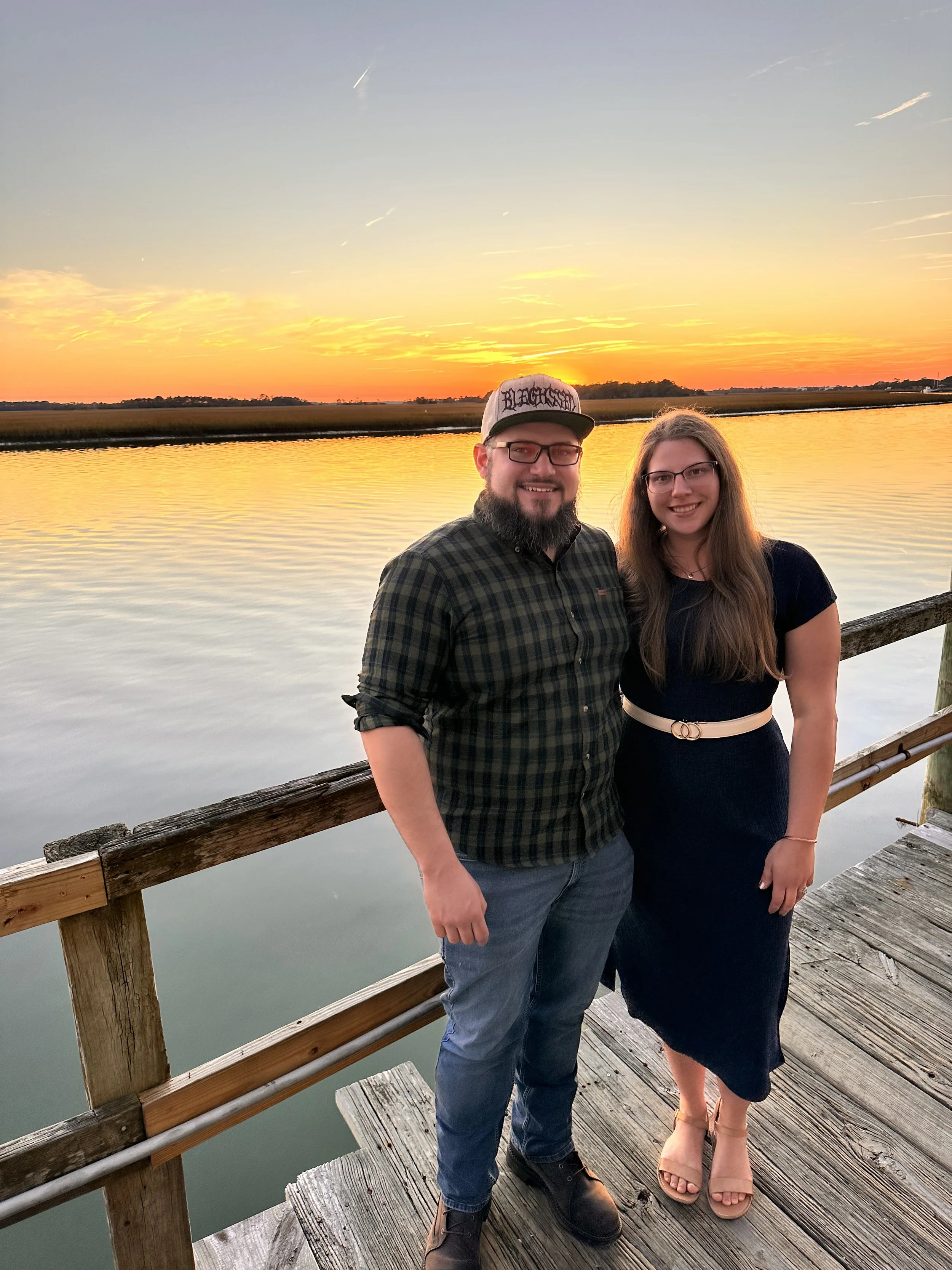 A man and woman standing on a wooden dock at sunset, with water in the background and a partly cloudy sky.