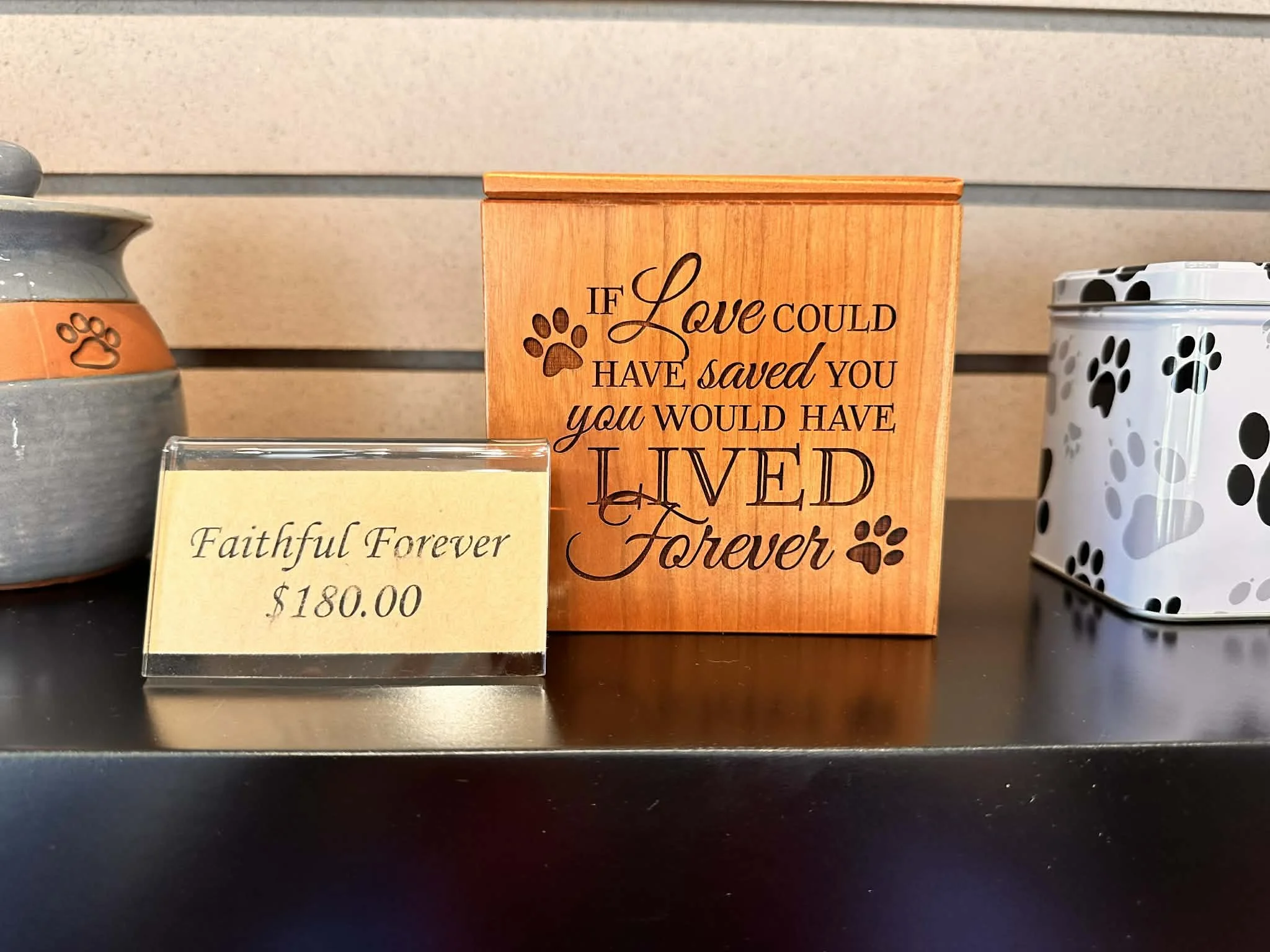 Decorative wooden sign with black text that reads: 'If Love Could Have Saved You, You Would Have Lived.' To the left of the sign is a ceramic object with an orange band featuring paw print designs. To the right of the sign is a white canister with bl