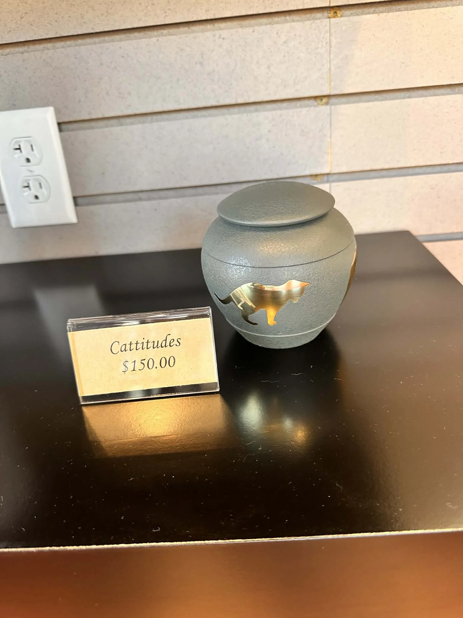 A gray decorative urn with a lid and cut-out design on a black surface, with a sign that reads 'Cattitudes $150.00'.