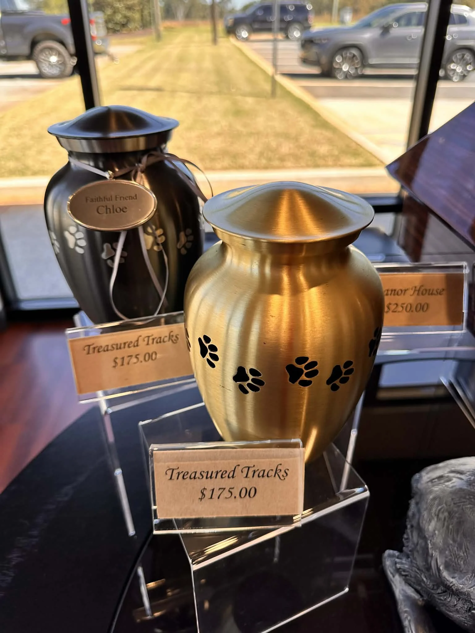 Decorative golden urn with black paw prints next to a black container and a sign that reads 'Treasured Tracks $175.00' displayed on a clear stand. The background features parked cars and a grassy area outside.