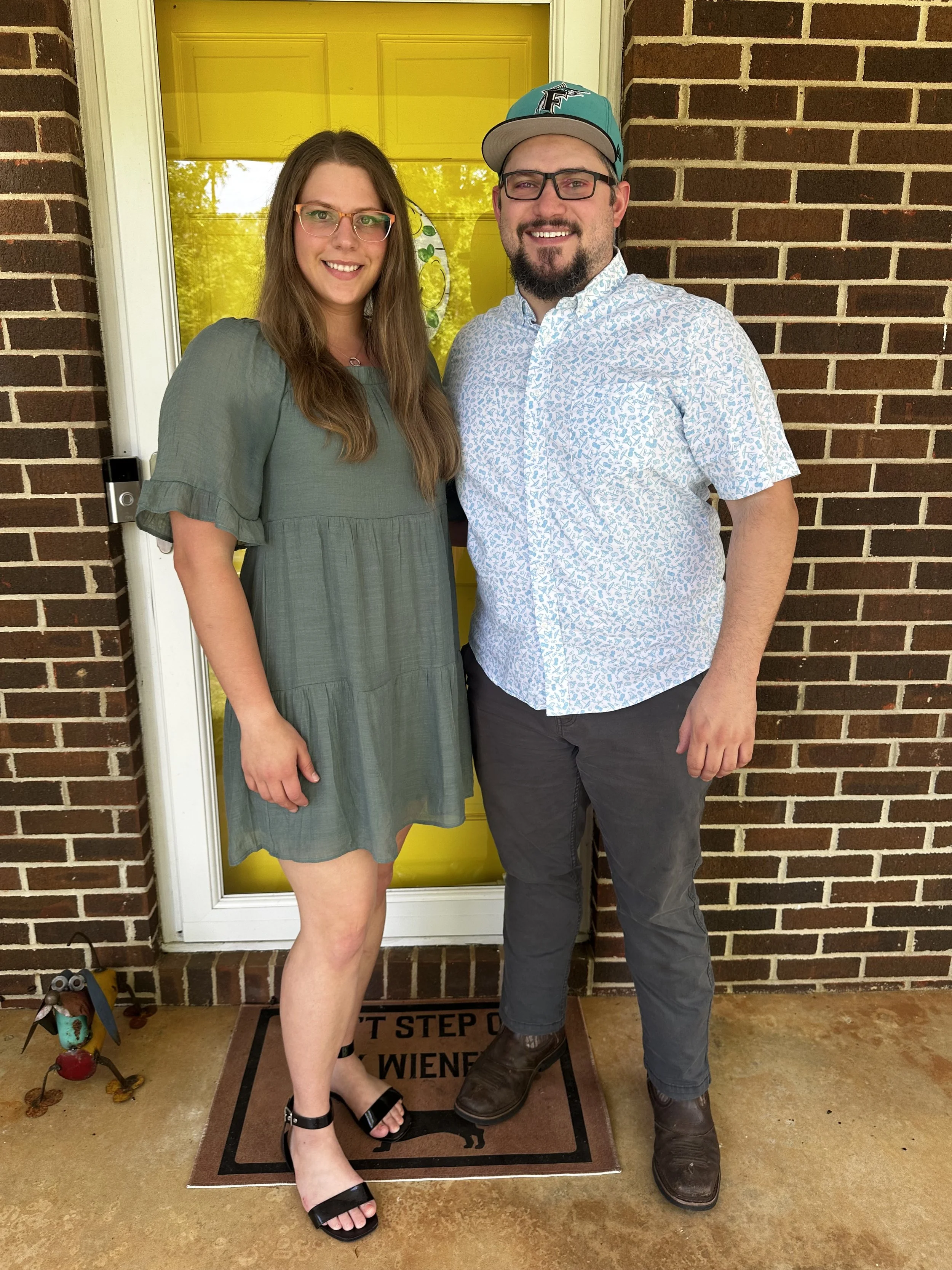 A smiling woman and man stand in front of a yellow door and brick wall, posing for a photo together. The woman has long brown hair, is wearing glasses, a green dress, and black sandals, while the man has a beard, glasses, a patterned shirt, dark pants, and cowboy boots, and is wearing a baseball cap.
