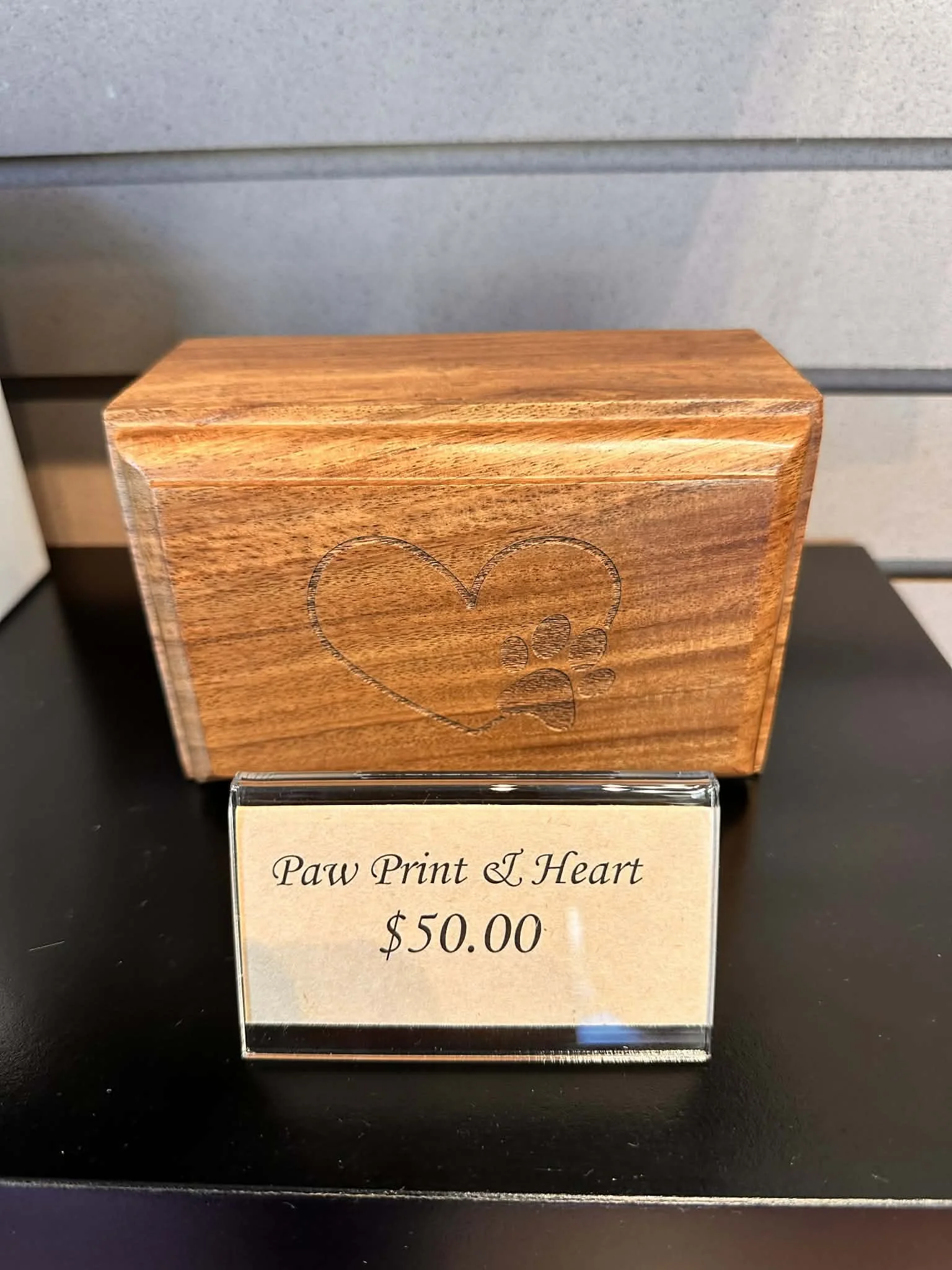 A wooden box with a paw print and heart engraving, displayed on a black surface, with a sign below indicating it is called 'Paw Print & Heart' priced at $50.00.
