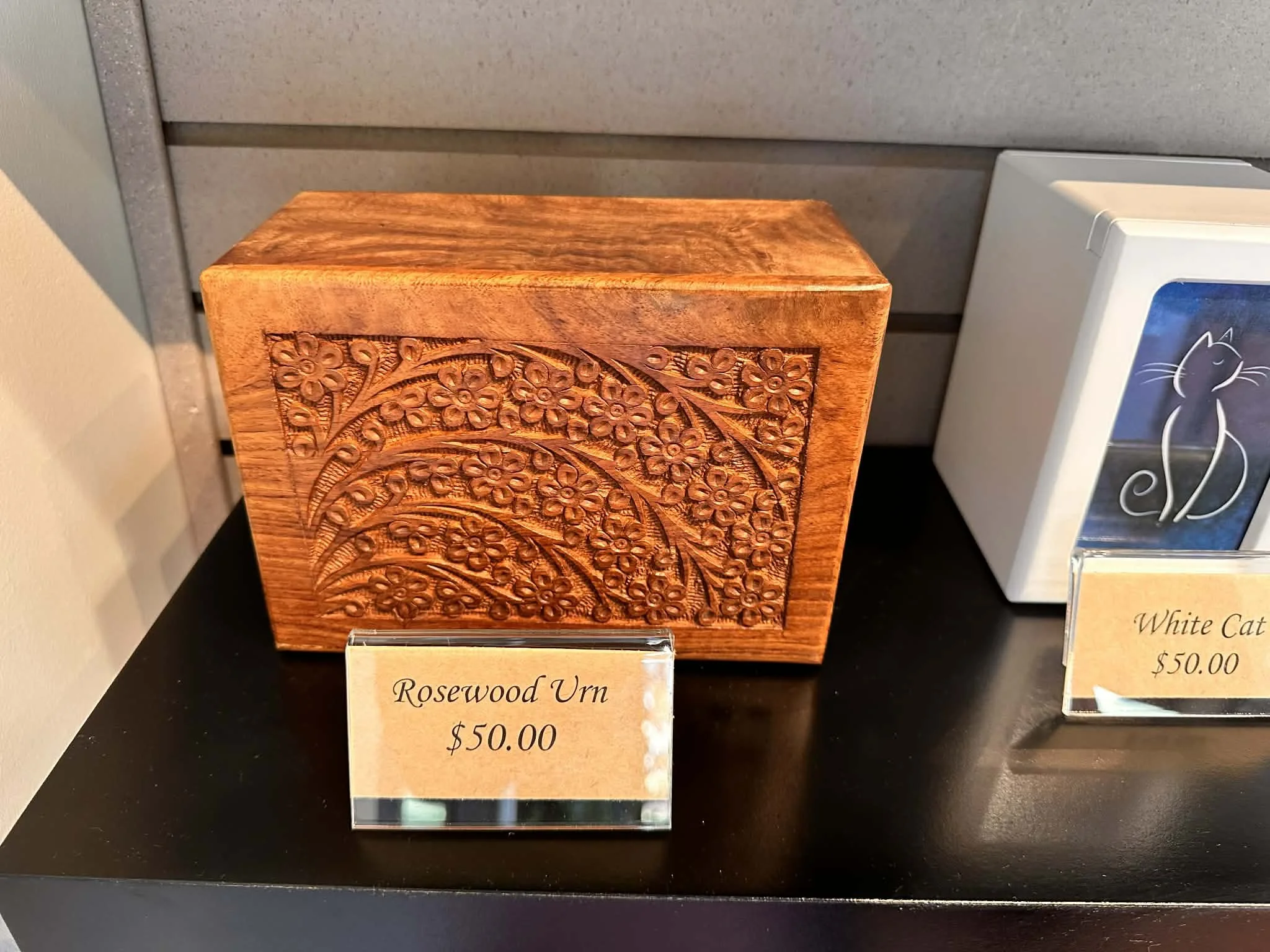 Decorative wooden urn with carved floral design, labeled 'Rosewood Urn' priced at $50.00, displayed beside a framed photograph of a black cat, priced at $50.00.