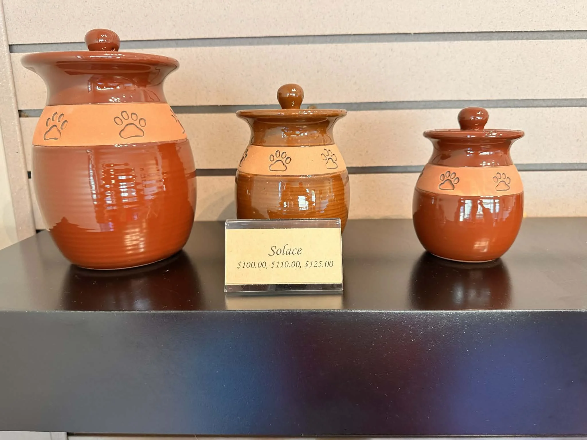 Three brown ceramic vases with paw print designs on a black surface, displayed in a row in front of a beige wall with horizontal blue stripes. A small yellow price tag reads "Solace $100.00, $110.00, $125.00."