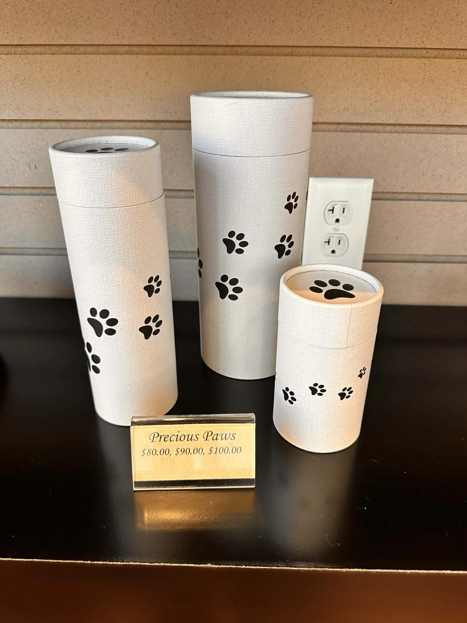 A display of three white cylindrical containers with black paw prints on them, labeled as 'Precious Paws'. The containers are priced at $80.00, $90.00, and $100.00. There is a small sign in front indicating the names and prices.