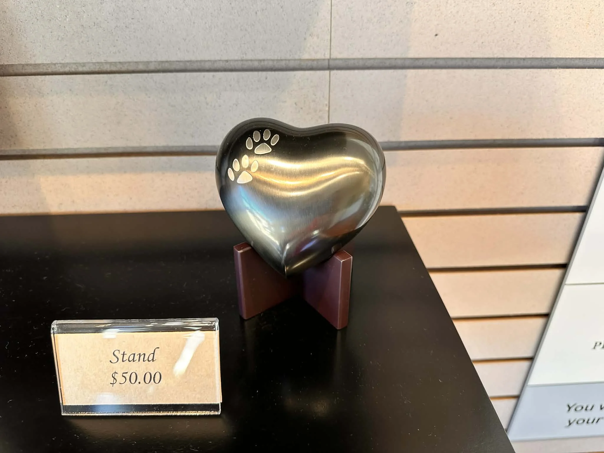 A metallic heart-shaped sculpture with paw print cutouts, placed on a stand on a black surface. A display sign nearby shows a price of $50.00.