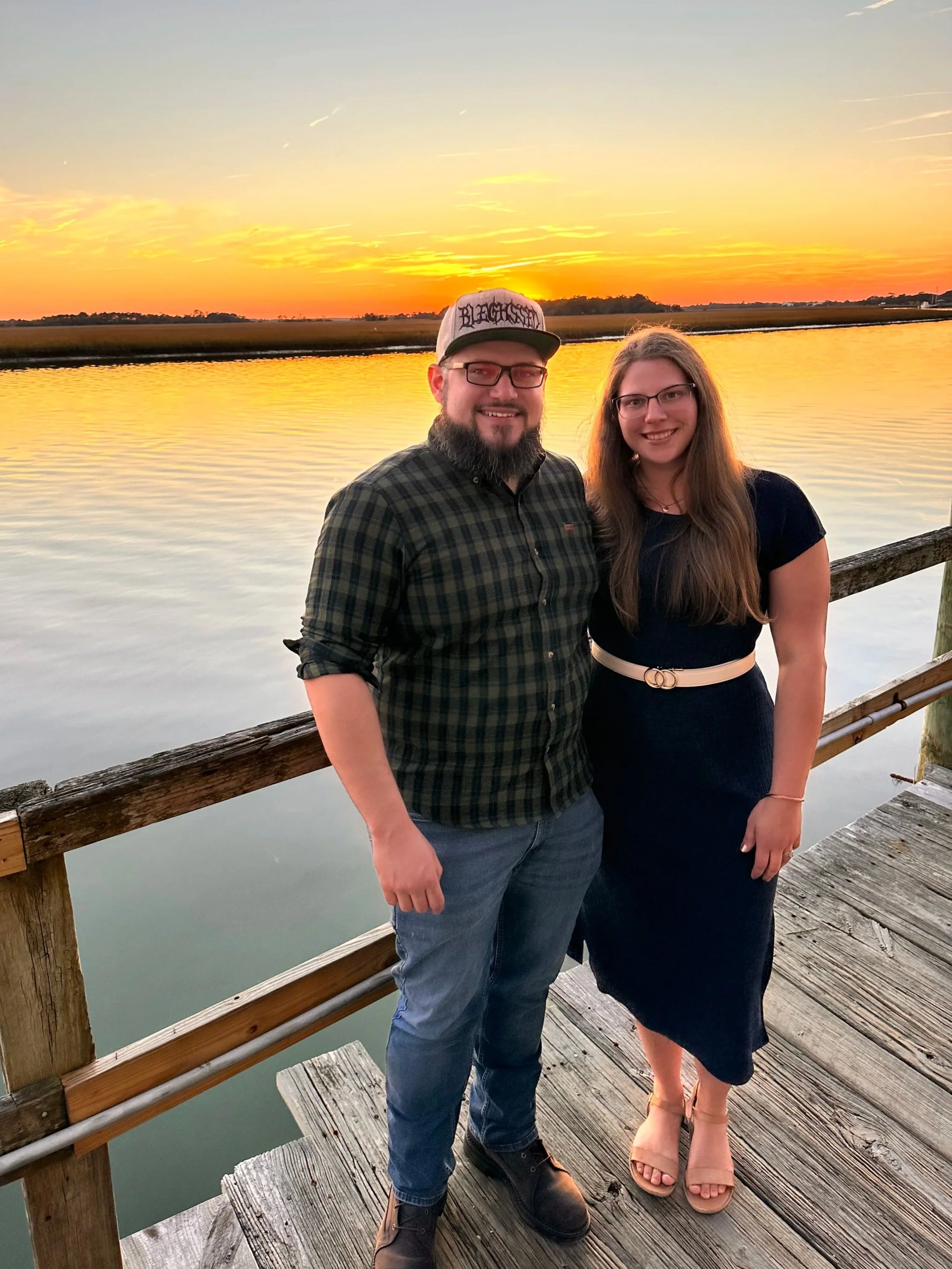 A man and woman stand on a wooden dock during sunset. The man is wearing a checked shirt, jeans, glasses, and a cap that says "Blackser". The woman is wearing a dark dress with a beige belt, and tan sandals. They are smiling at the camera with water and a colorful sunset sky in the background.