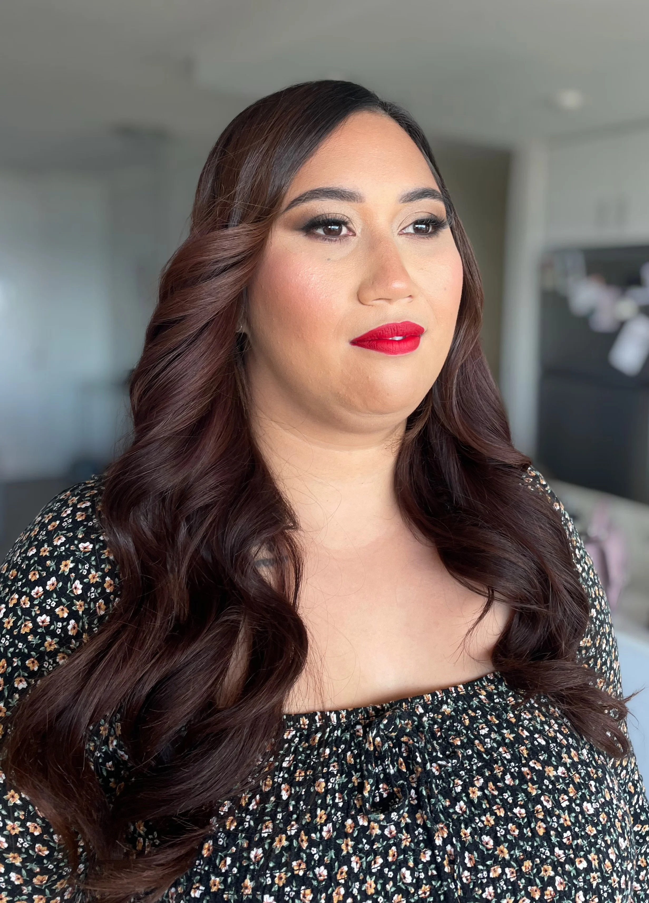 A woman with long wavy brown hair styled with loose curls, wearing makeup with red lipstick, smoky eye makeup, and a floral black dress with a square neckline.