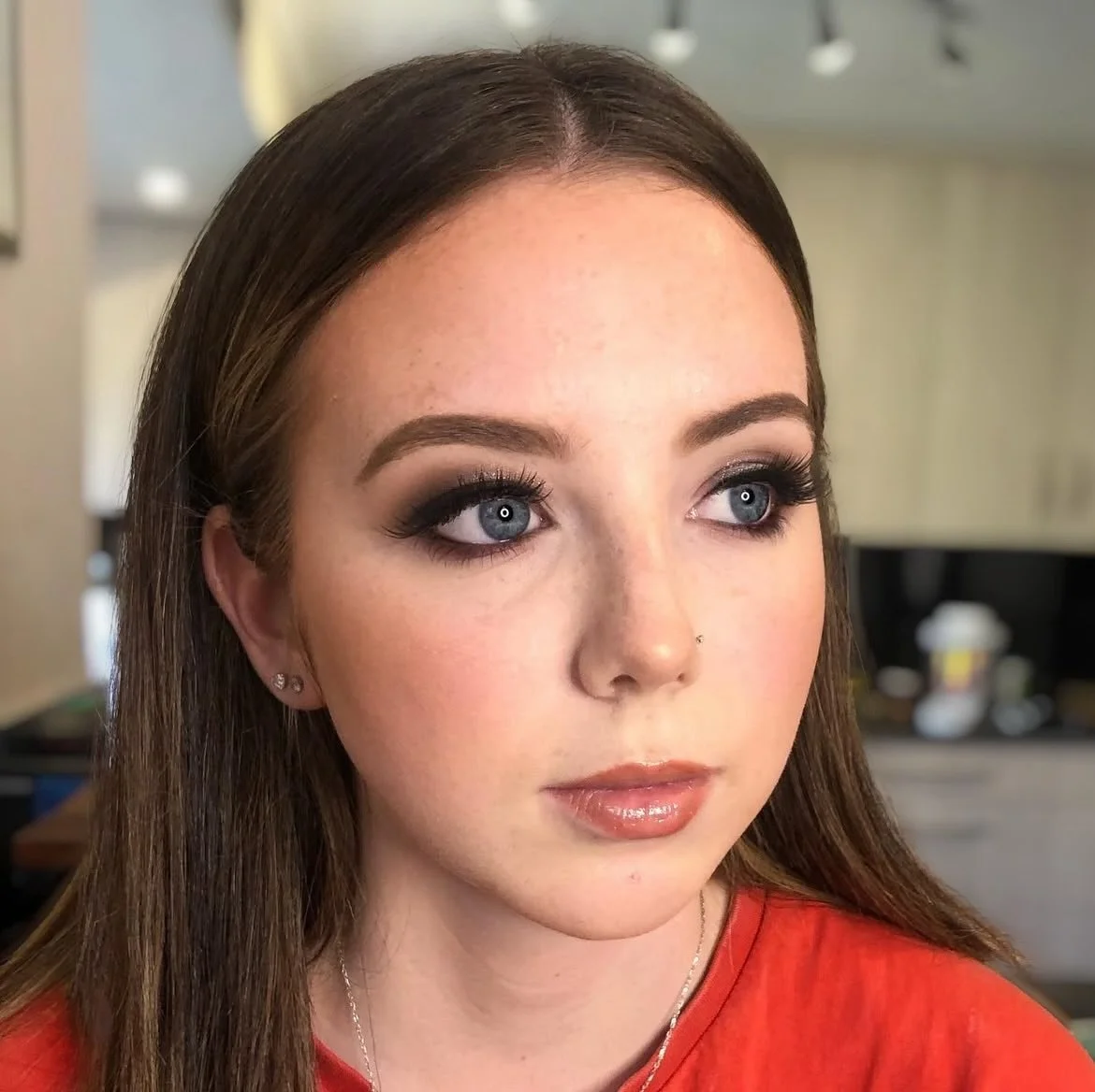 Close-up of a young woman with long brown hair, blue eyes, and makeup, wearing a red top and earrings, in an indoor setting.