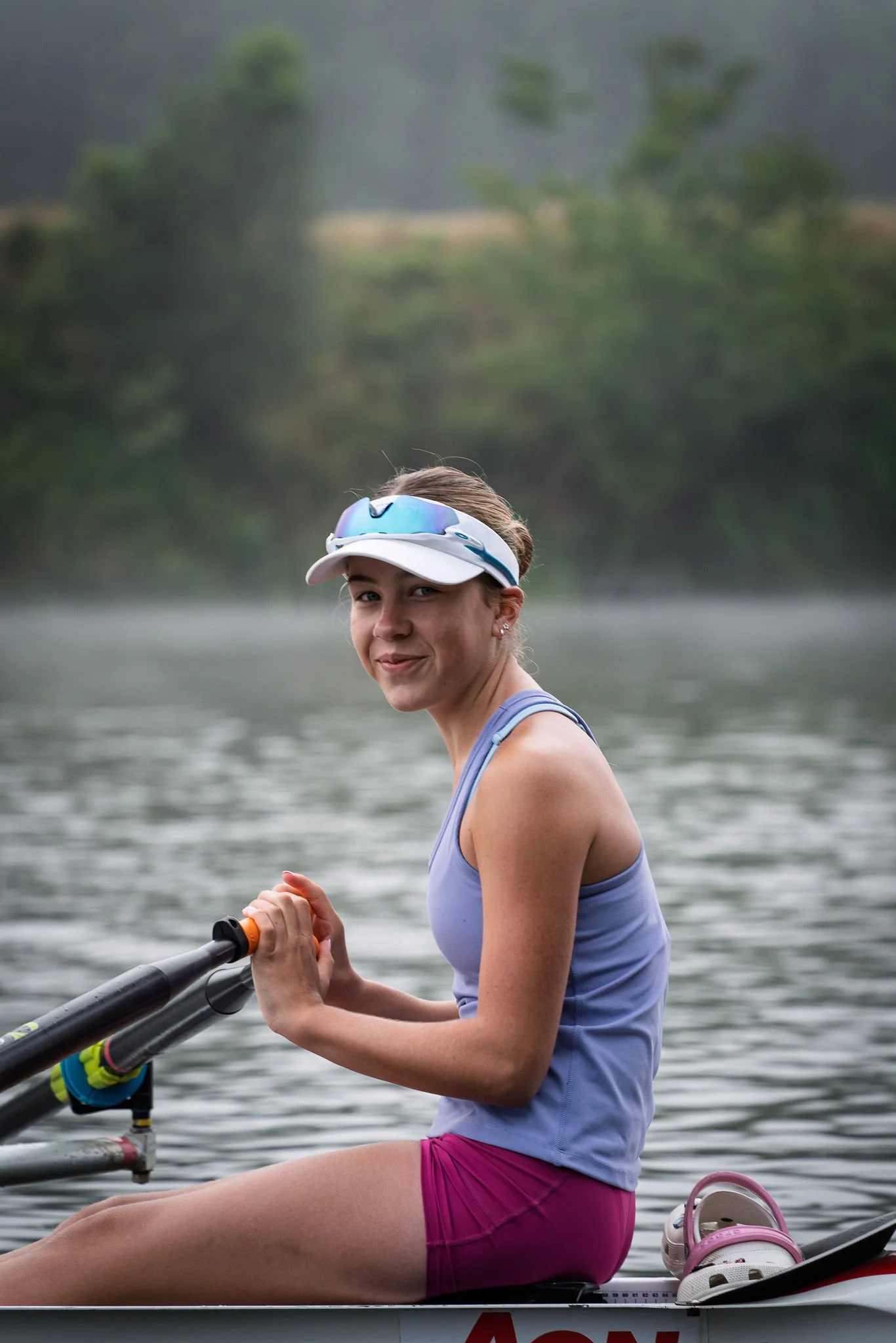 A young woman sitting in a kayak on a river, wearing a blue tank top, pink shorts, a white and blue visor, and holding a paddle, with a scenic river and trees in the background.