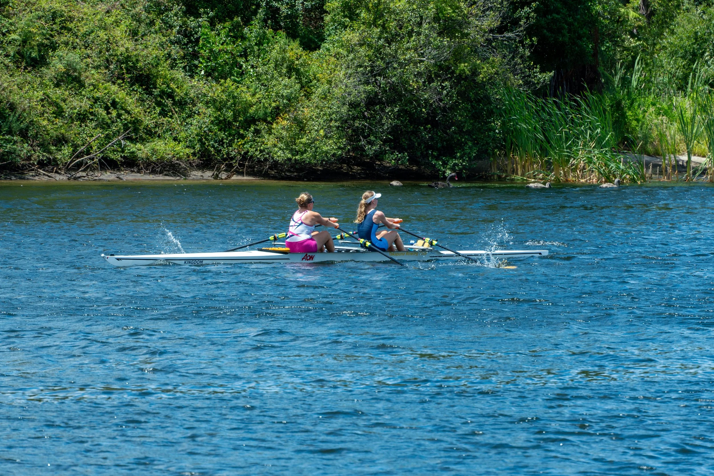 Two women in a rowing boat on a river with green trees and bushes in the background.