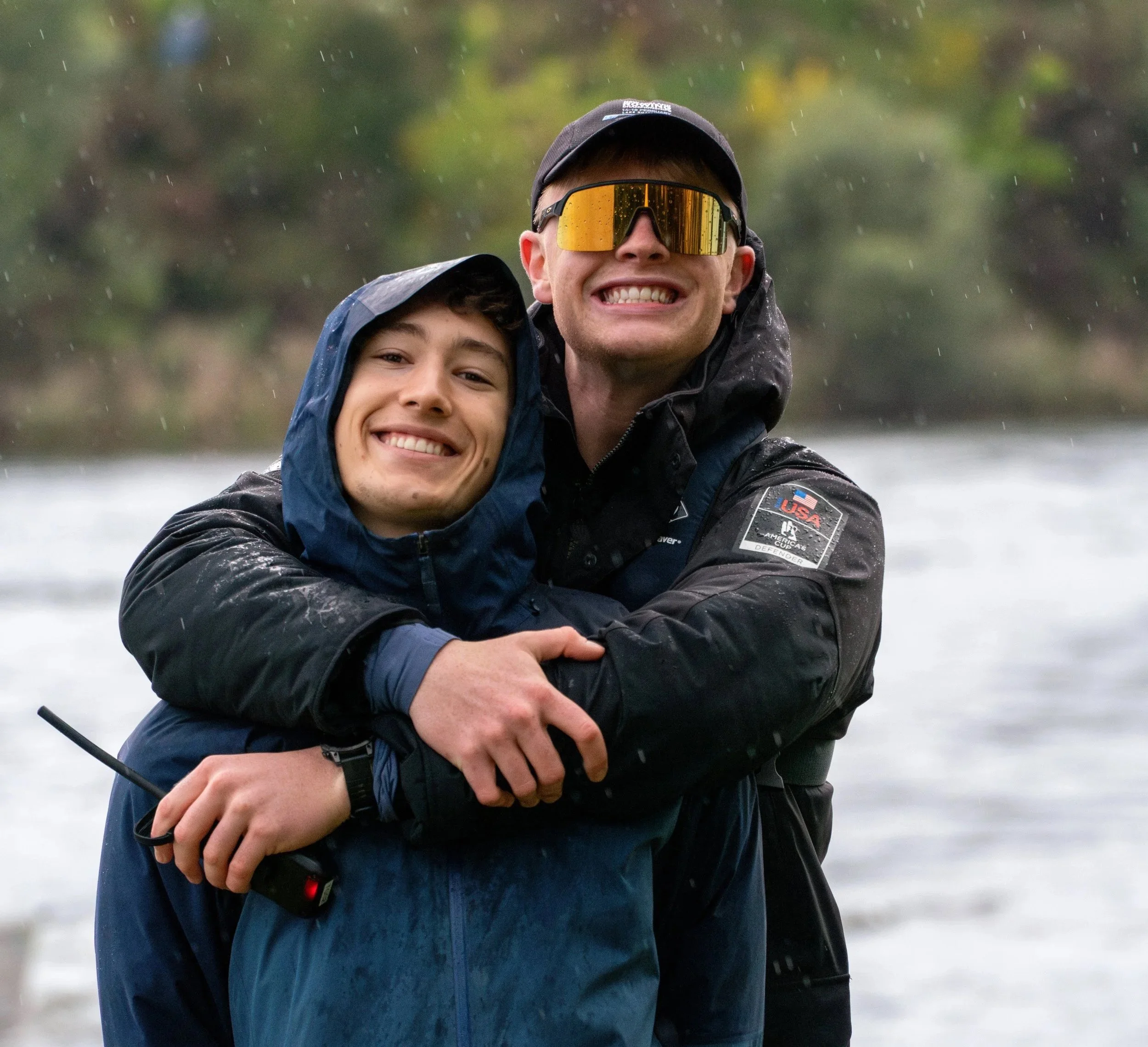 Two smiling men hugging outdoors near a body of water, wearing rain jackets and waterproof gear, with a blurred green landscape in the background.