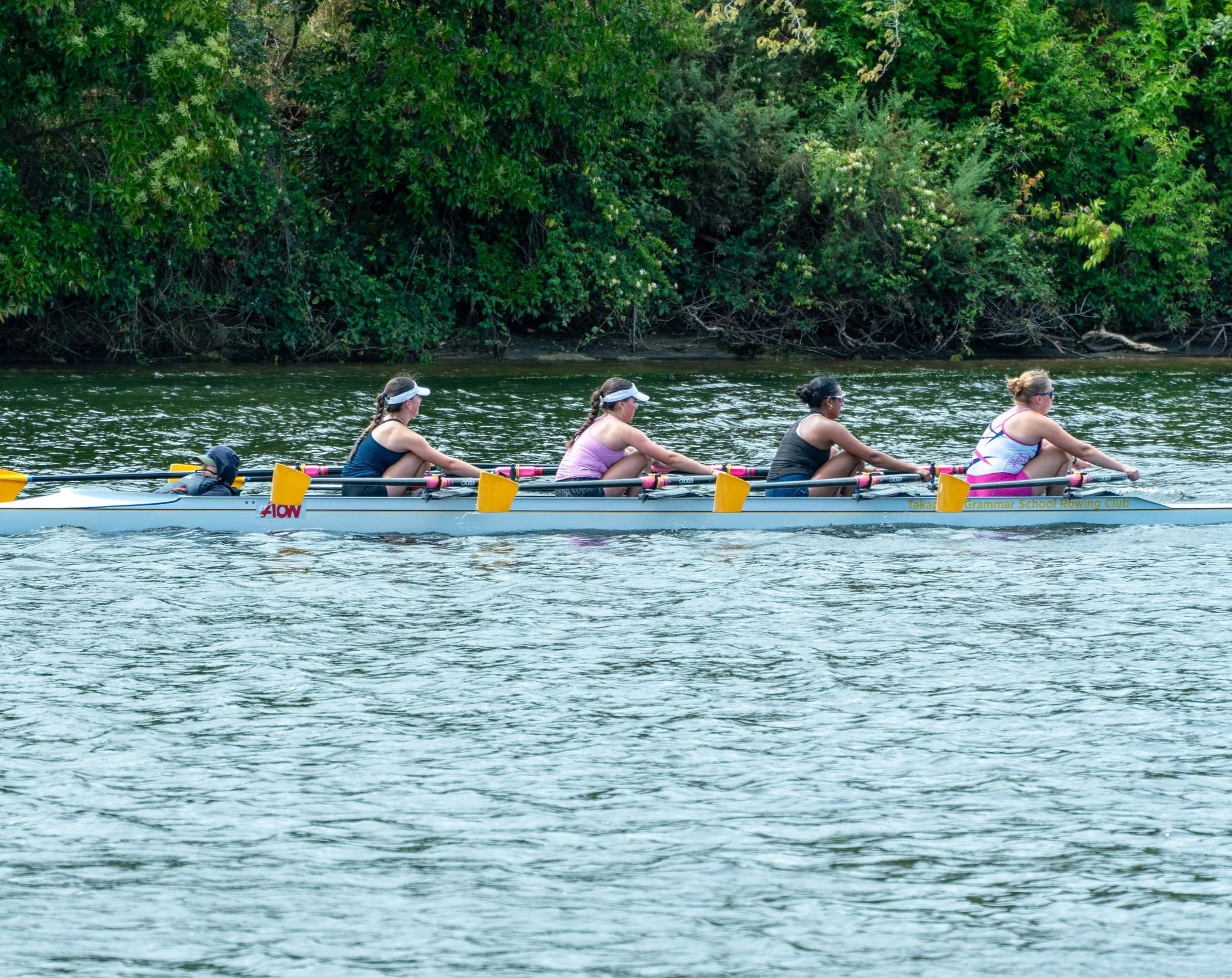 A team of five women rowing a racing shell on a river, with dense green trees in the background.