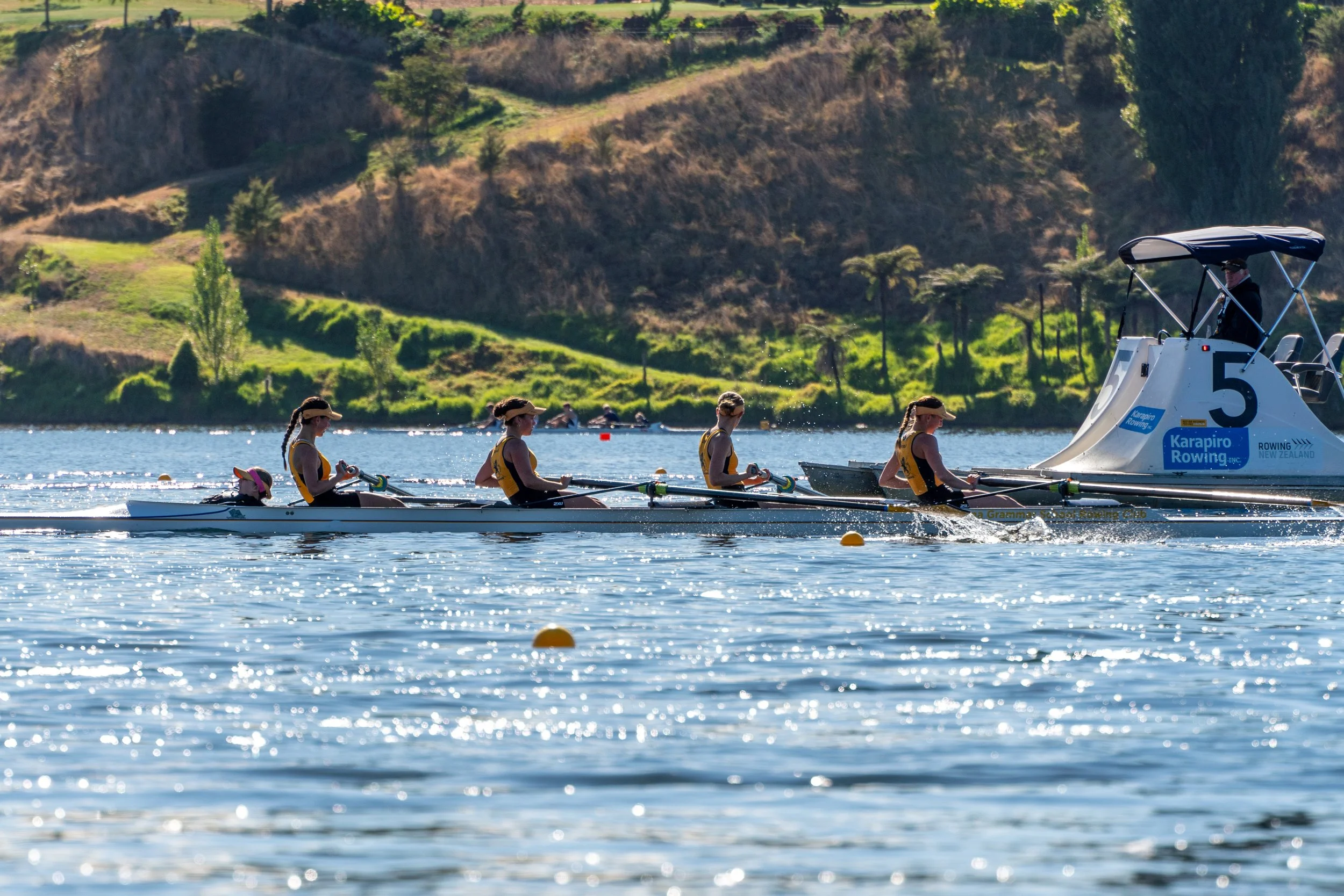 Four female rowers in a racing boat on a lake, being coached by a person in a motorboat with a blue and white logo, and a large number 5. The background features grassy hills and trees.