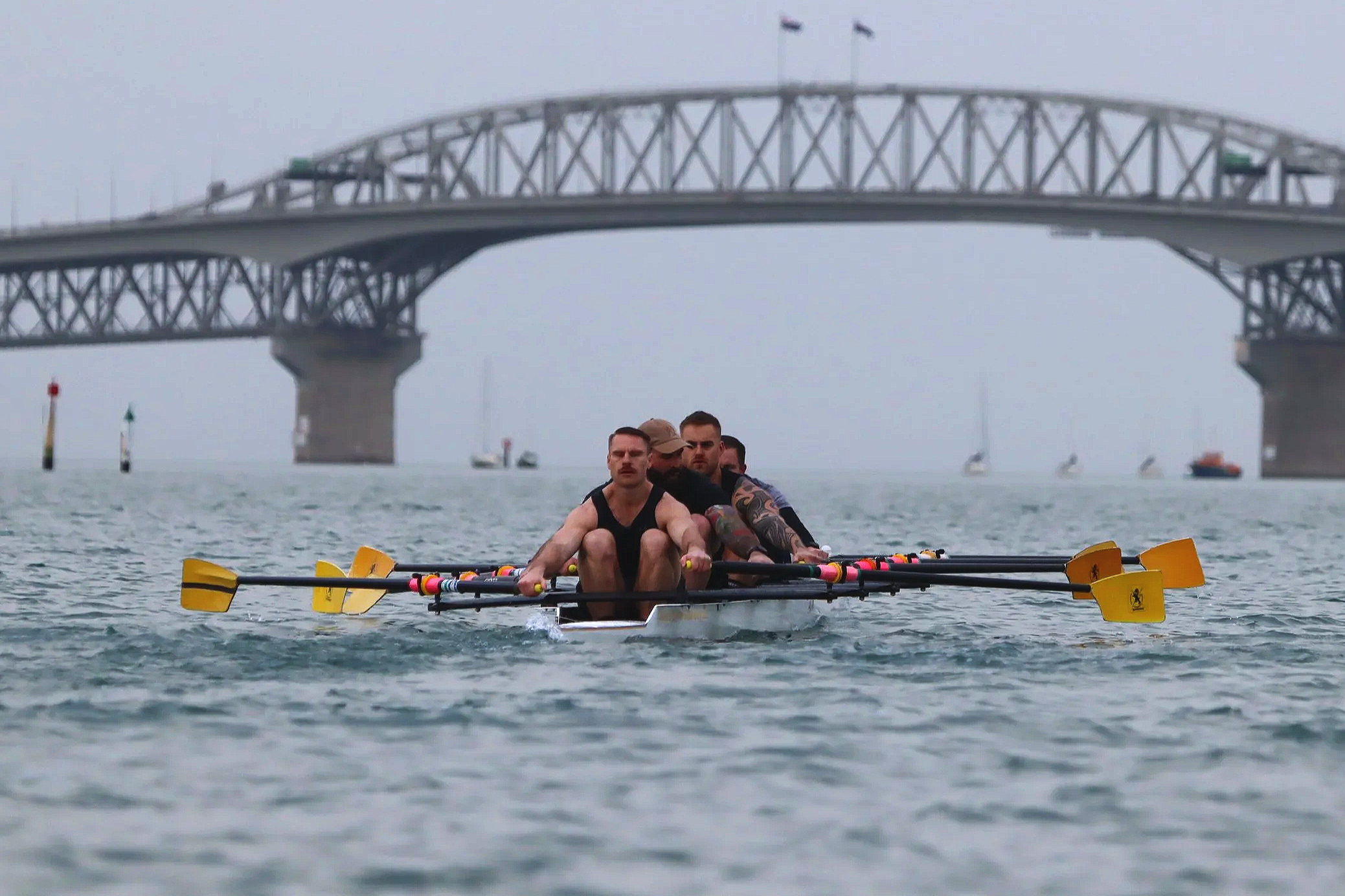 Four men rowing a boat on the water under a large bridge with sailboats in the background.
