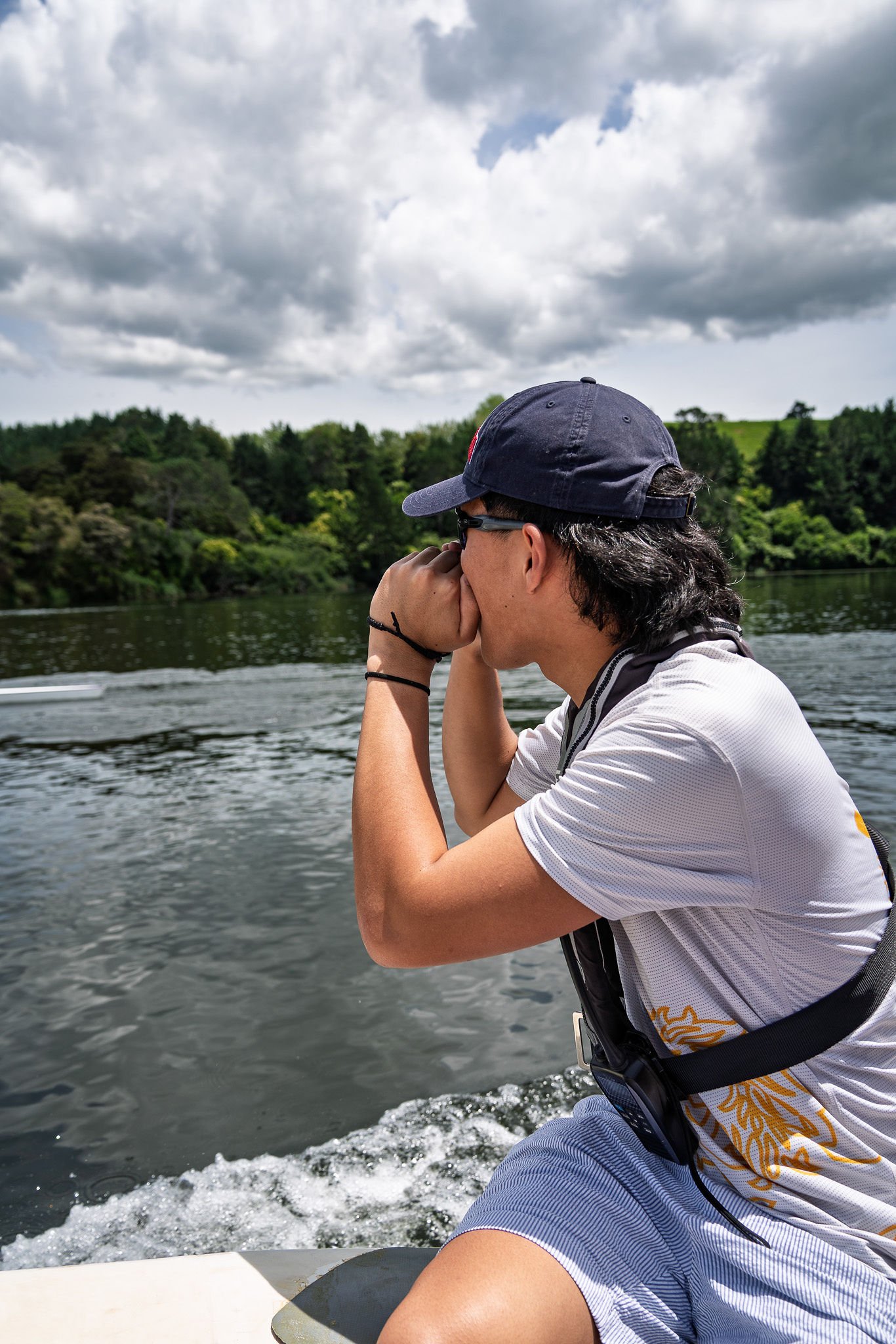 A man in a white shirt and blue cap looking through binoculars while on a boat on a river, with a background of trees and cloudy sky.