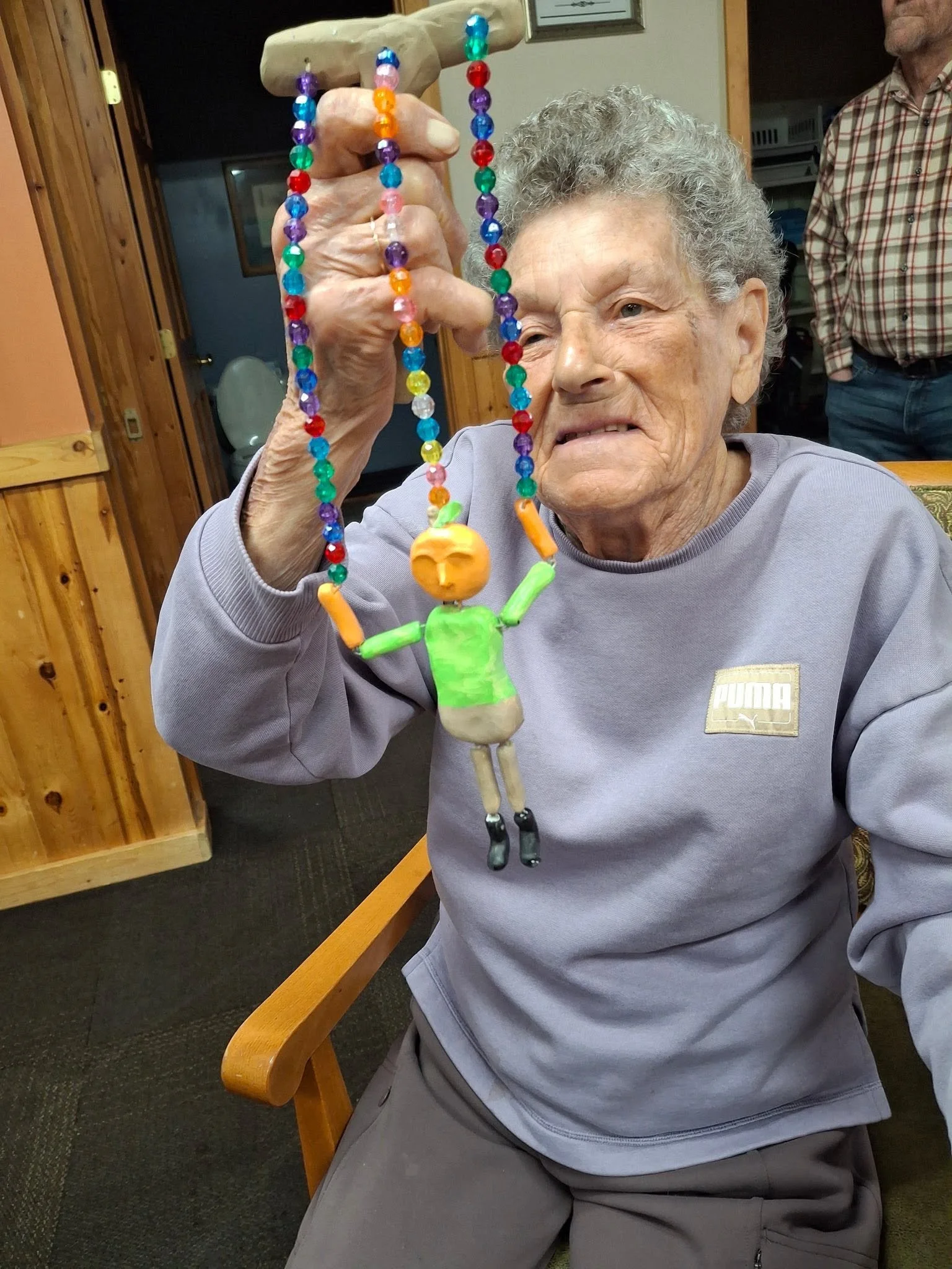 An elderly woman sitting at a wooden table, holding up a colorful beaded necklace with a string of brightly colored beads and a small doll figure hanging at the bottom. She appears to be examining the necklace thoughtfully.